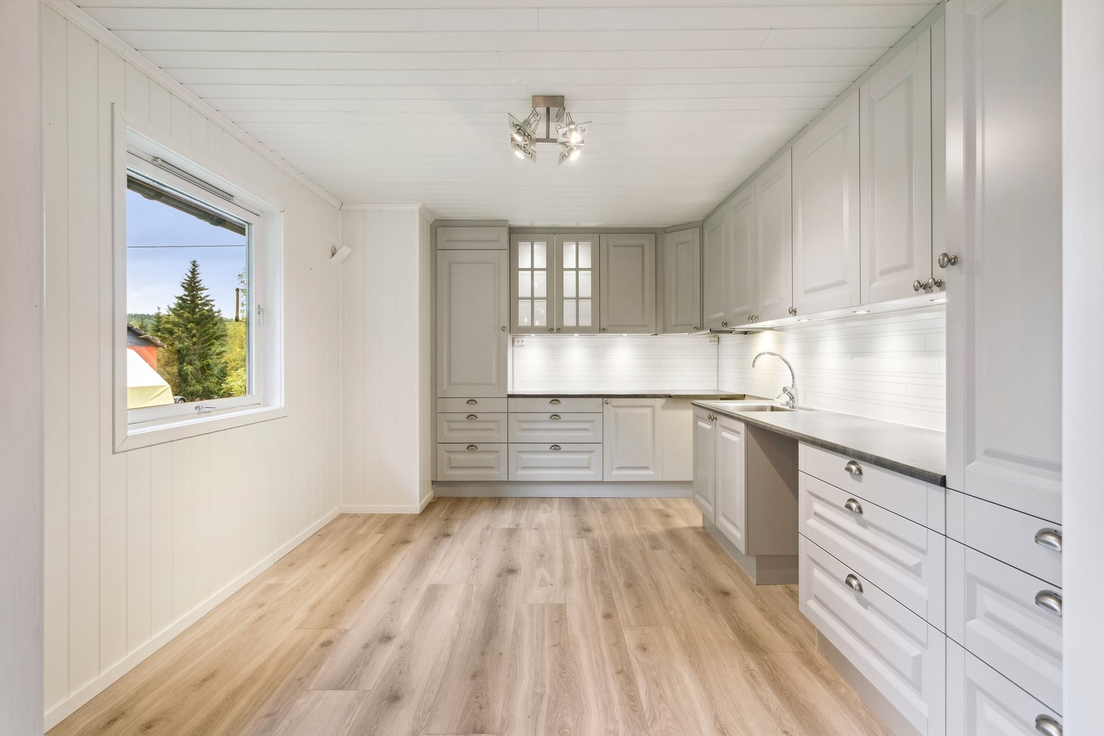 Kjøkkenet har hvitmalt stående panel på veggene og lys laminat på gulvet. Lys grå kjøkkeninnredning i hjørneløsning med laminerte skrog, formpressede fronter og laminerte benkeplater. Galleribilde