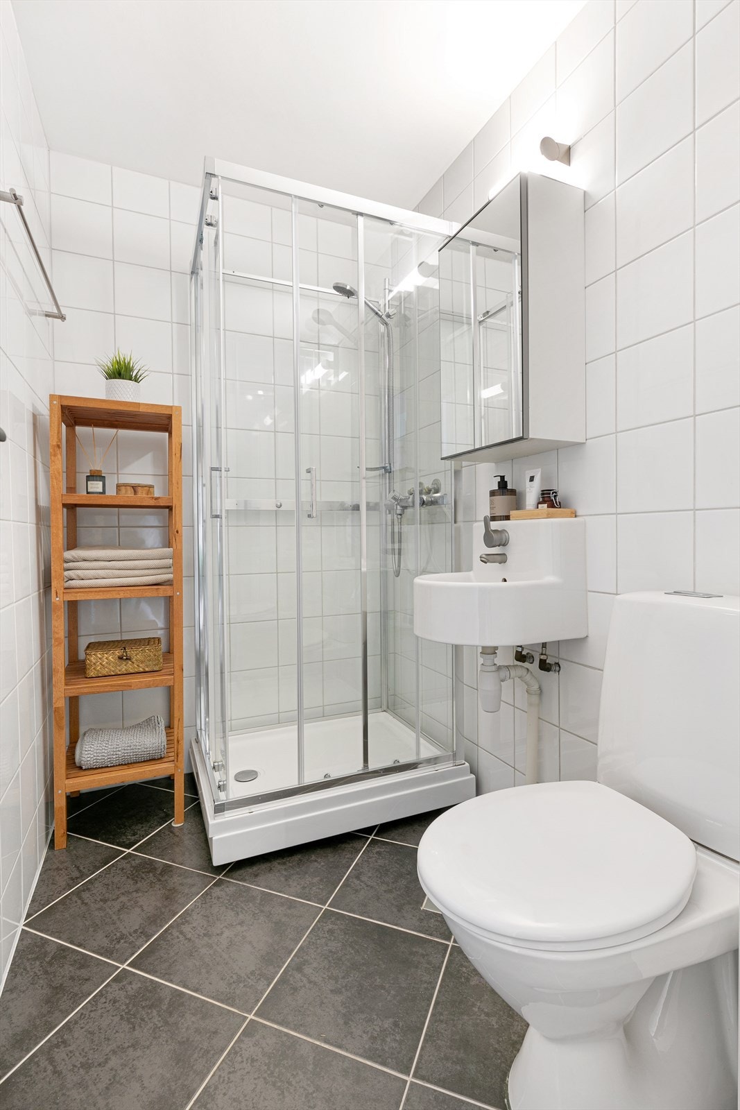 Flislagt baderom med alt dere trenger! Badet inneholder baderomsinnredning med glatte, folierte fronter, servant og speilskap montert på vegg og gulvmontert toalett (2017). I hjørnet av rommet er det dusjkabinett fra 2023 med skyvedører og dusjutstyr. Galleribilde