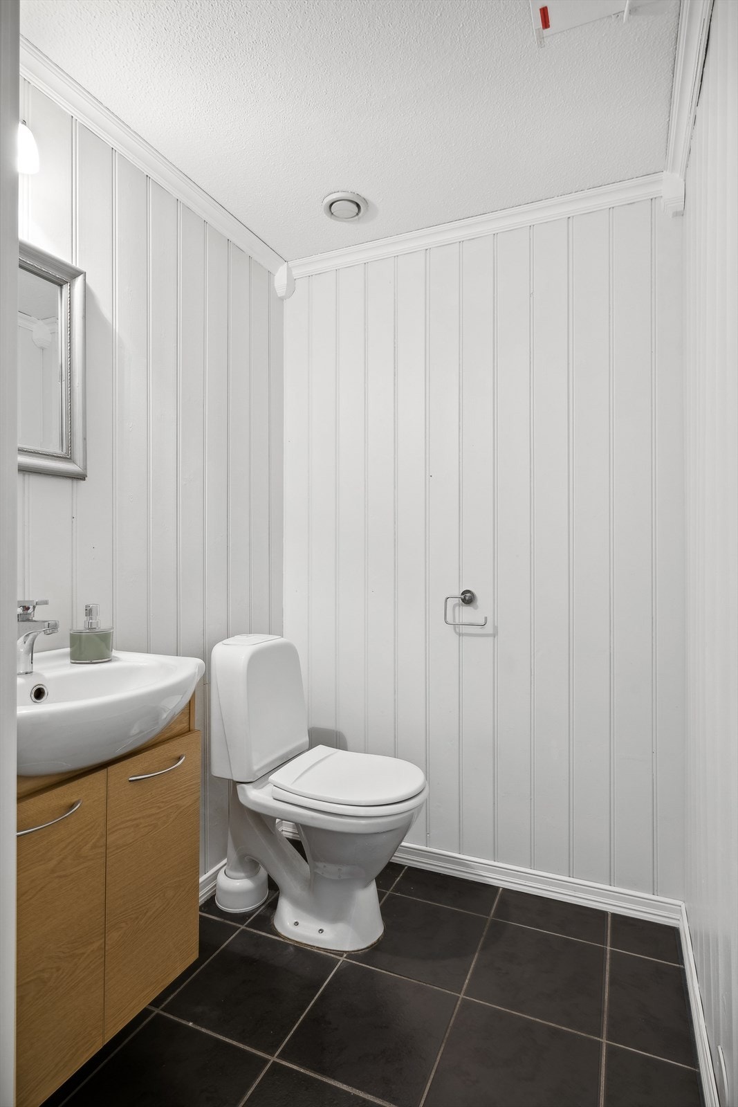 Toalettrom med fliser på gulv og vegger med malte plater. Rommet har innredning med servant med skapseksjon, speil og gulvstående toalett. Galleribilde