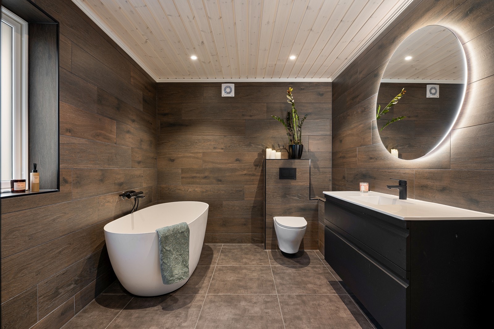 Meget pent bad fra 2023 med flislagt gulv med gulvvarme og mørke fliser i treimitasjon på veggoverflatene. Galleribilde