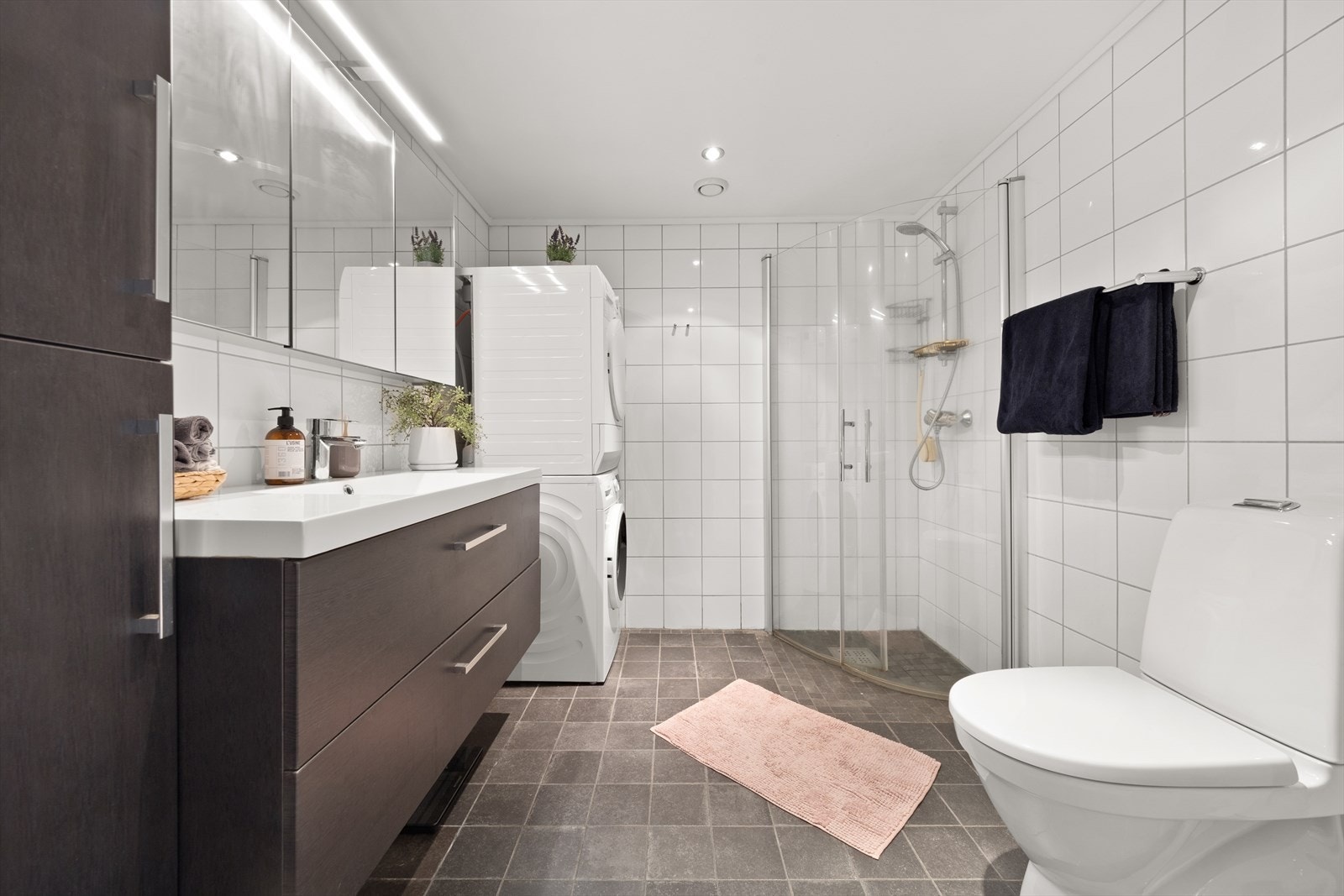 Pent flislagt bad med innredning bestående klosett, dusjhjørne med innfellbare dører og baderomnsinnredning med speilskap. Badet har opplegg for vaskemaskin og tørketrommel Galleribilde