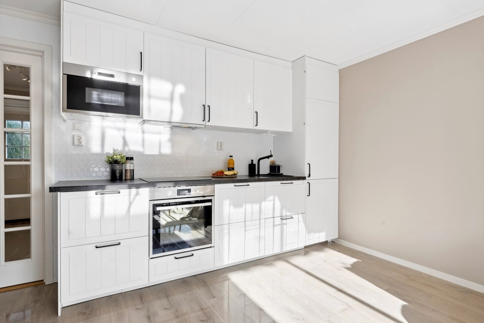 Delikat IKEA kjøkkeninnredning fra 2023 profilerte fronter, belegg på vegg over benkeskap. Laminat benkeplate med nedfelt kum. Ventilator. Induksjonstopp, stekeovn, oppvaskmaskin, mikrobølgeovn og kjøl/frys er integrert i innredningen. Galleribilde