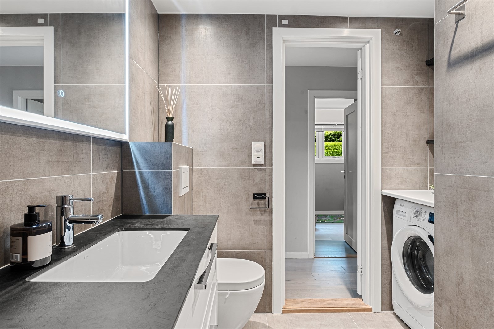 Badet er innredet med dusjhjørne, servant med underskap og speil og veggmontert toalett. Praktisk benk med opplegg for vask/tørk. Galleribilde
