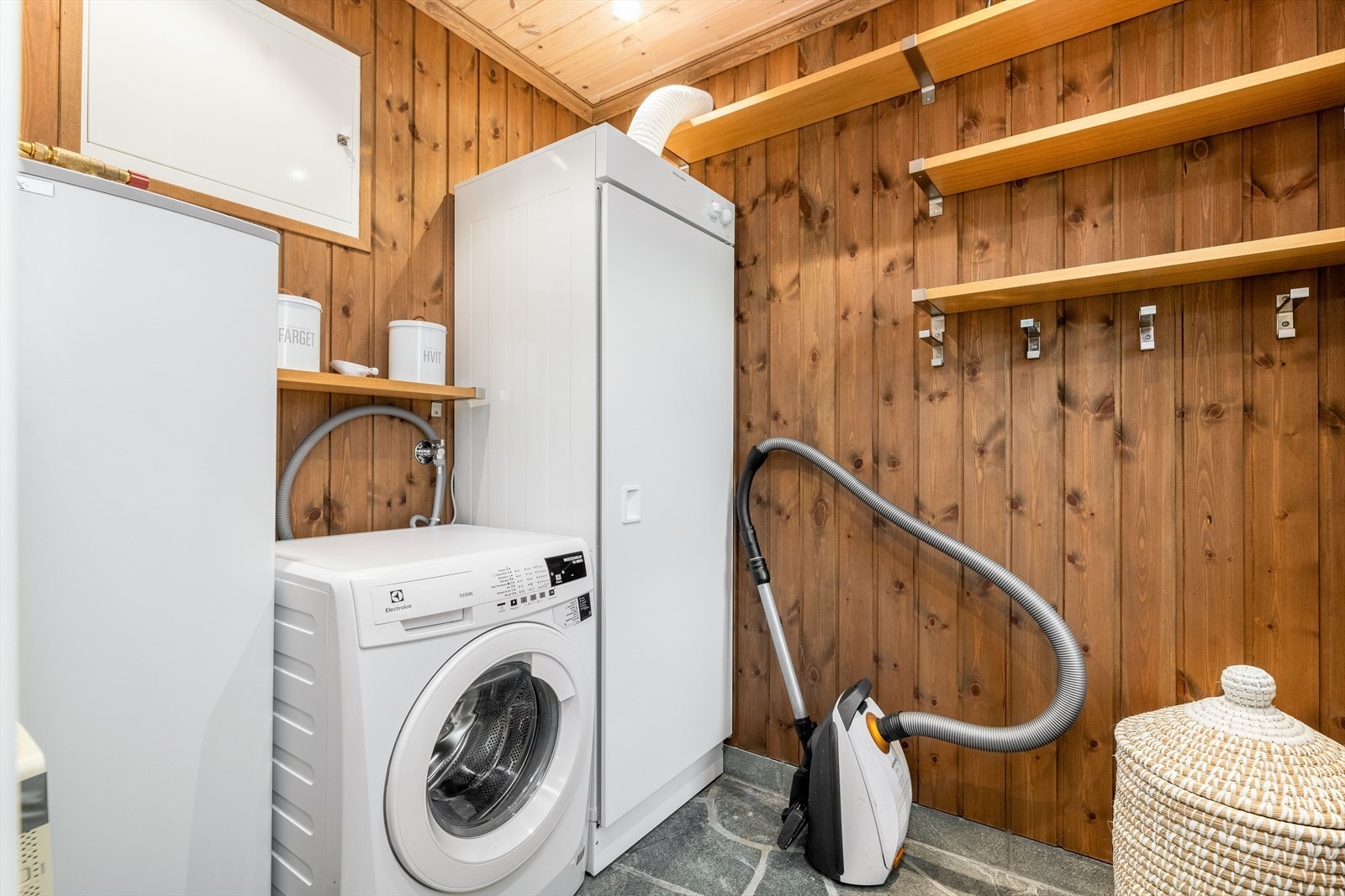 Romslig vaskerom med panel på vegger og i himling samt skifer på gulv. Her er det mye hylleplass til oppbevaring samt knagger til henge fra seg yttertøy. Vaskerommet er utstyrt med opplegg for vaskemaskin og en varmtvannstank på 160 liter. Galleribilde