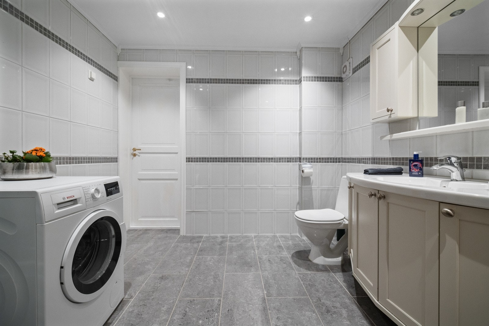 Rommet har veggfliser med fliseborder av kontrastfargede mosaikkfliser og rektangulære gulvfliser over gulvvarme. Himling er pent lyssatt med downlights og det er vegghengt toalett Galleribilde