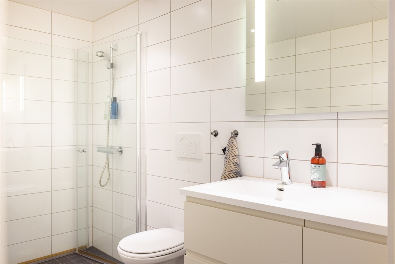 Badet er innredet med vegghengt toalett, servant med innredning og dusjhjørne med innfellbare glassdører. Galleribilde