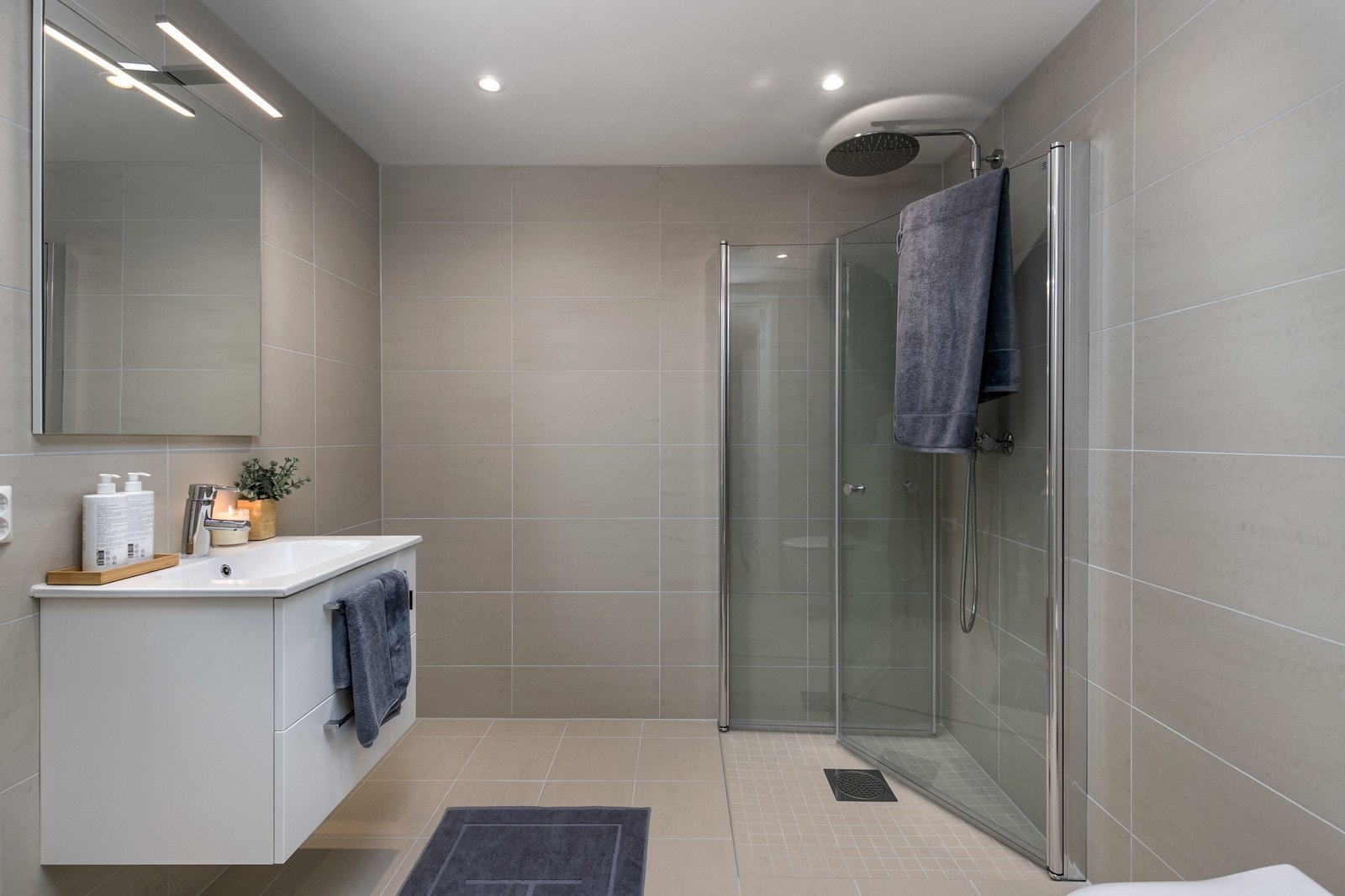 Delikat og plassbygd baderom med sandfargede fliser, downlights i tak, samt hvit innredning med servant og speil. Innfellbare dusjvegger og opplegg til vaskemaskin. Galleribilde