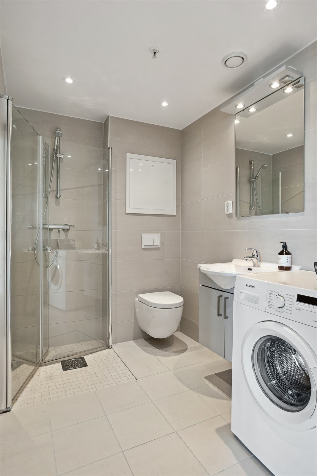 Badet inneholder dusjhjørne med innfellbare glassdører, innredning med servant, vegghengt toalett og opplegg for vaskemaskin. Galleribilde