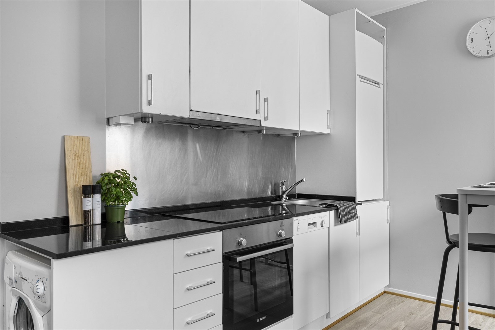 Kjøkkeninnredningen består videre av steinbenkeplate med nedfelt kum i rustfritt stål, stålplate på vegg, integrert induksjonsplatetopp og stekeovn, oppvaskmaskin og vaskemaskin i nisje og kjøle-/fryseskap er plassert i skap på kjøkken. Galleribilde