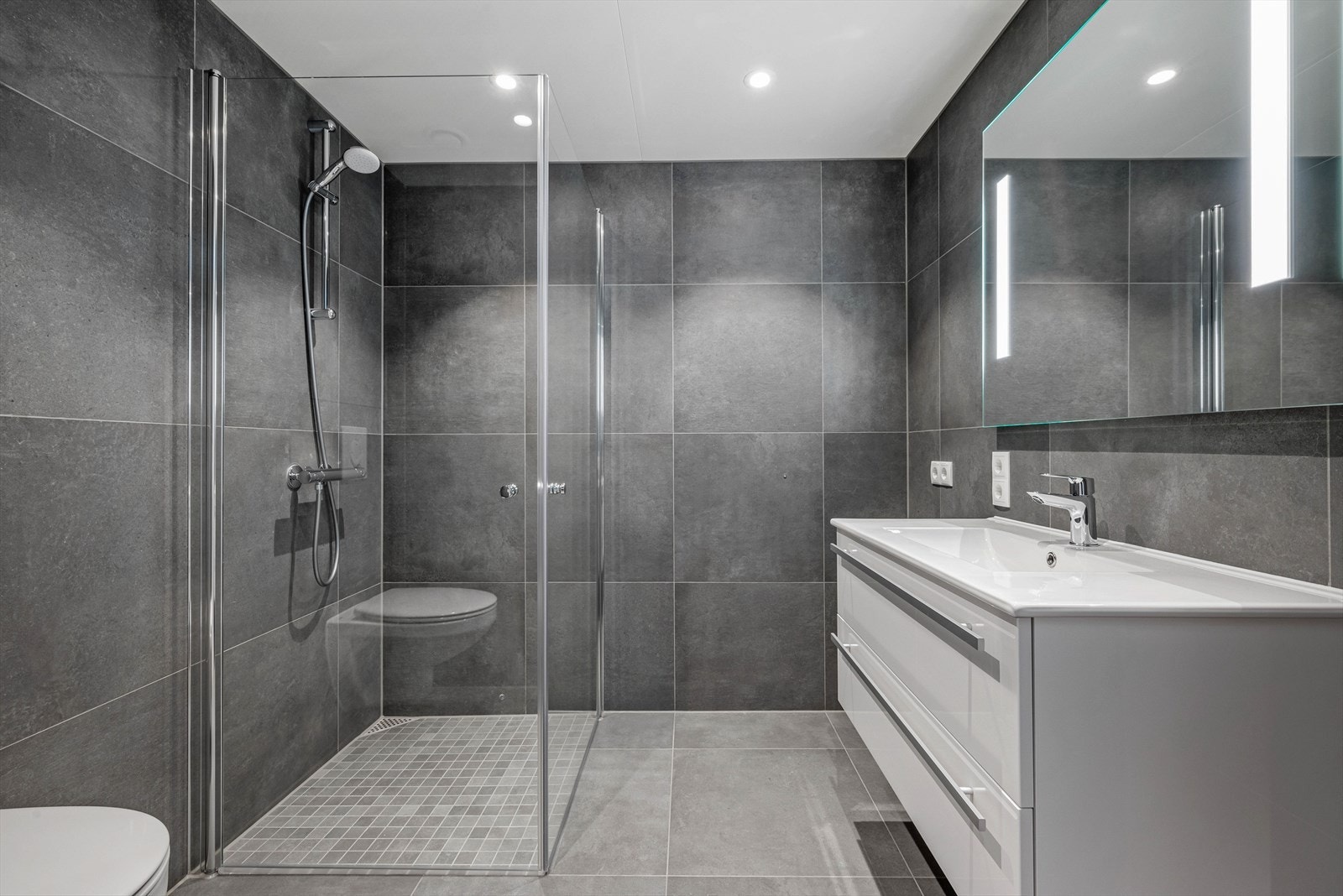 Badet har spotter i tak med dimming og nydelige fliser på gulv og vegger. Det er uttak til vaskemaskin ved baderomsinnredningen. Galleribilde