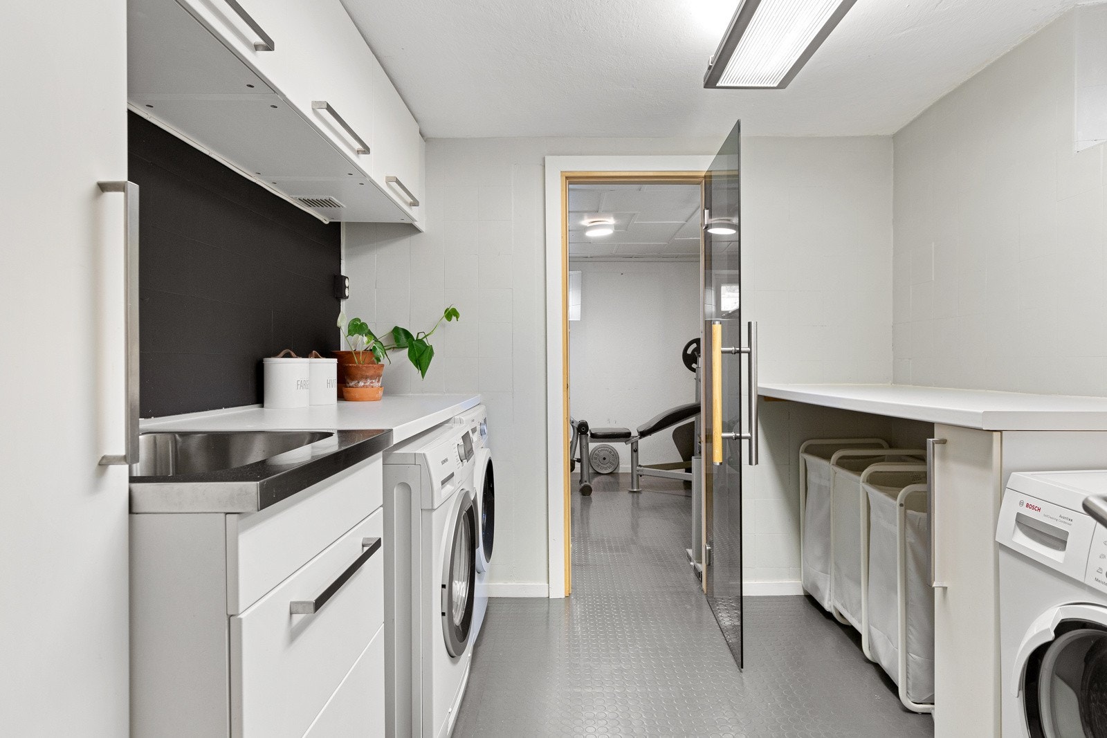 Vaskerom med industribelegg på gulv og malte fliser på veggene. Innredning har glatte fronter, laminat benkeplate og skyllekum i rustfritt stål. Her er det godt med plass til både vaskemaskin og tørketrommel. Galleribilde