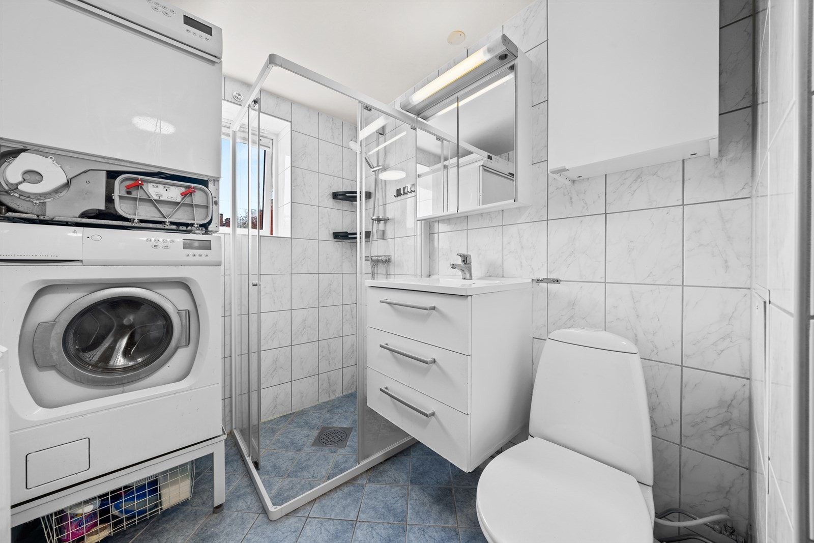 Baderommet er utstyrt med glatte, folierte fronter og heldekkende servant, speilskap med lysarmatur på vegg over servanten, opplegg for vaskemaskin og tørketrommel, gulvmontert toalett, dusjhjørne med skyvedører, sluk i sone og dusjutstyr. Galleribilde