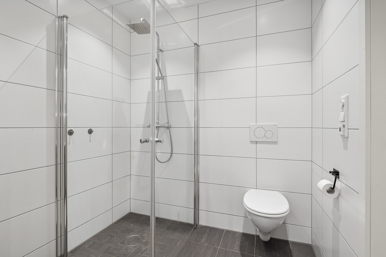 Badet er innredet med vegghengt toalett, samt et stort dusjhjørne med innfellbare glassdører, nedsenket gulv og dusjgarnityr med hånddusj og tropedusj montert på vegg. Galleribilde