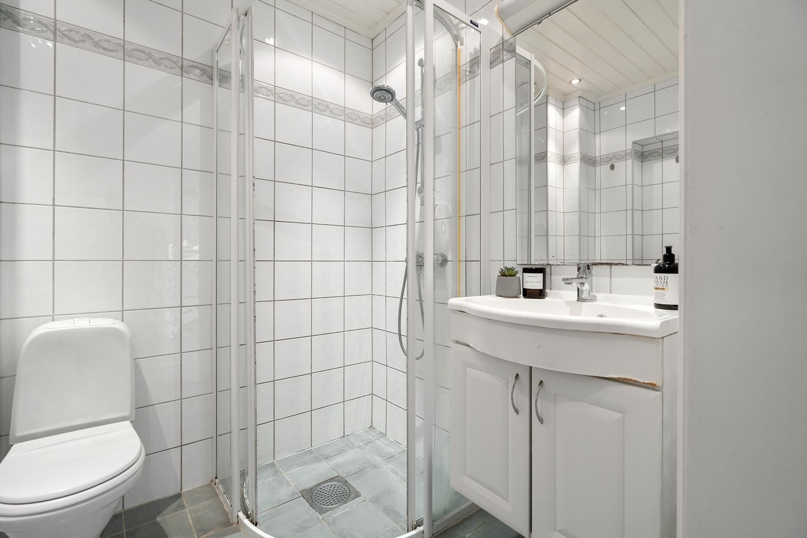Badet har dusjhjørne med fastmontert gulvskinne med skyvedører, gulvmontert toalett, servantskap med overmontert hvit servant og veggmontert speil på vegg. Galleribilde