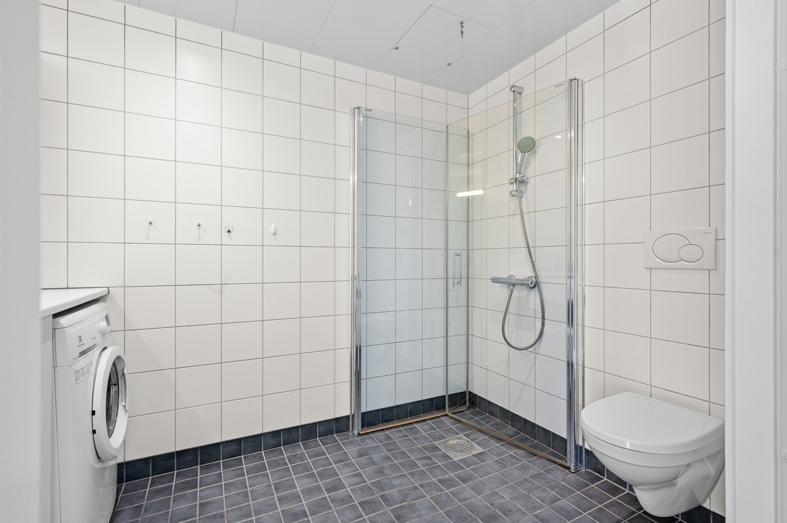 Baderom fra byggeår med flislagte overflater, varme i gulv og downlights i himlingen. Innredningen består av vegghengt servantskap med glatte fronter, benkeplate av laminat og nedsenket servant, vegghengt speil med overbelysning og stikkontakt. Galleribilde