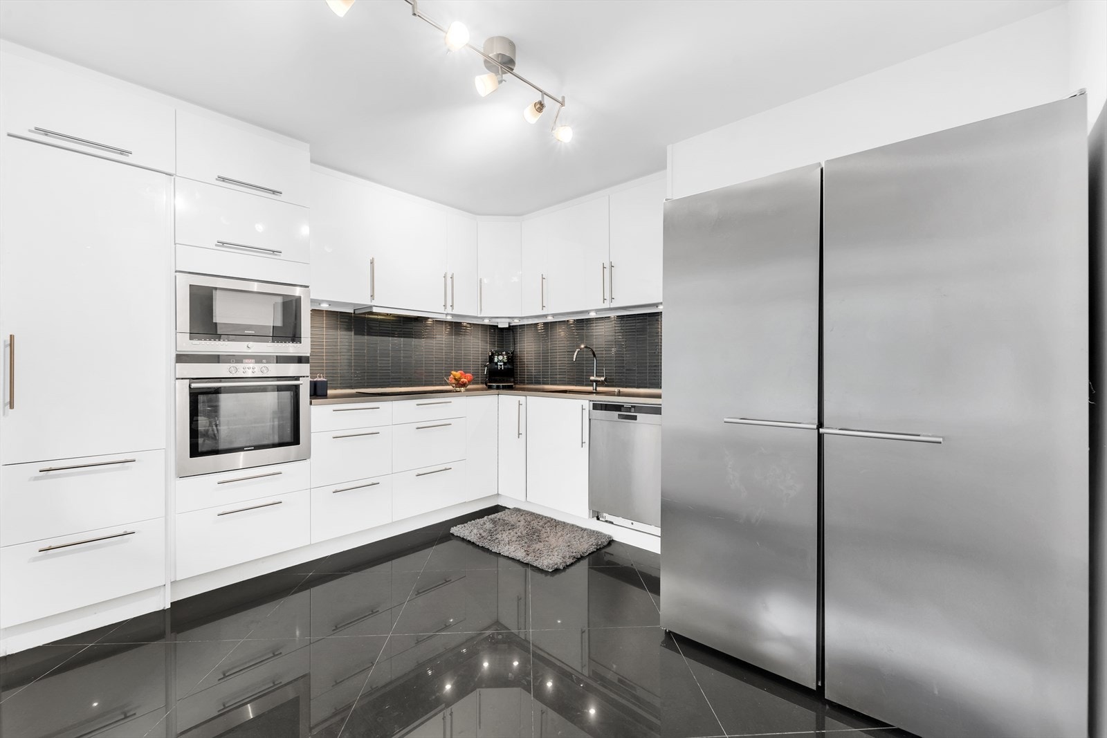 Kjøkkeninnredning med slette høyglans fronter, laminat benkeplate med fliser over, enkel underlimt oppvaskkum, uttrekksventilator, opplegg for oppvaskmaskin og integrerte hvitevarer som stekeovn, koketopp og mikroovn. Oppvaskmaskin medfølger. Galleribilde