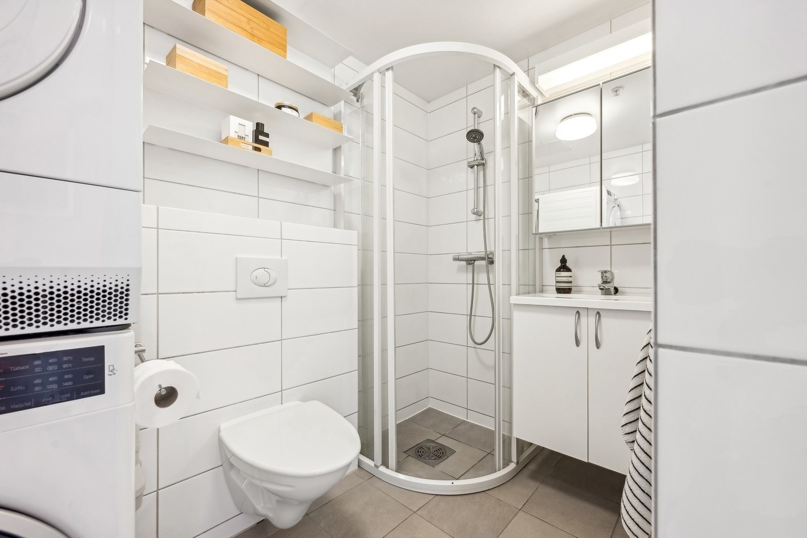 Badet er helfliset og pent, og det er montert elektriske varmekabler i gulvet. Rommet har innredning med nedfelt servant, veggmontert toalett, dusjvegger/hjørne og opplegg for vaskemaskin. Galleribilde