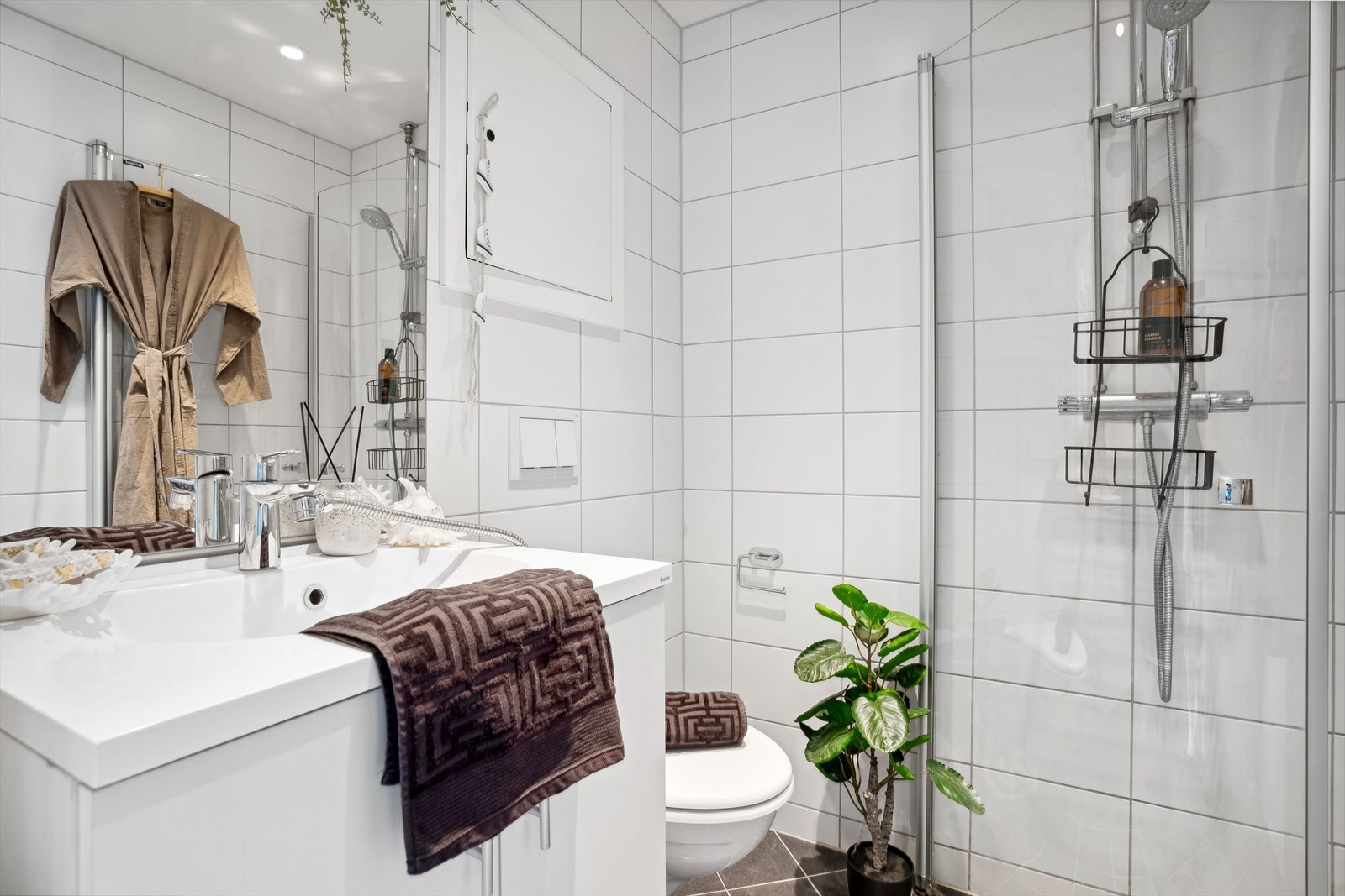 Baderommet er innredet med buet glass dusjvegg, vegghengt toalett med skjult sisterne, servant med skap under og ettgreps kran, speil på vegg med lys og stikkontakt over. Galleribilde