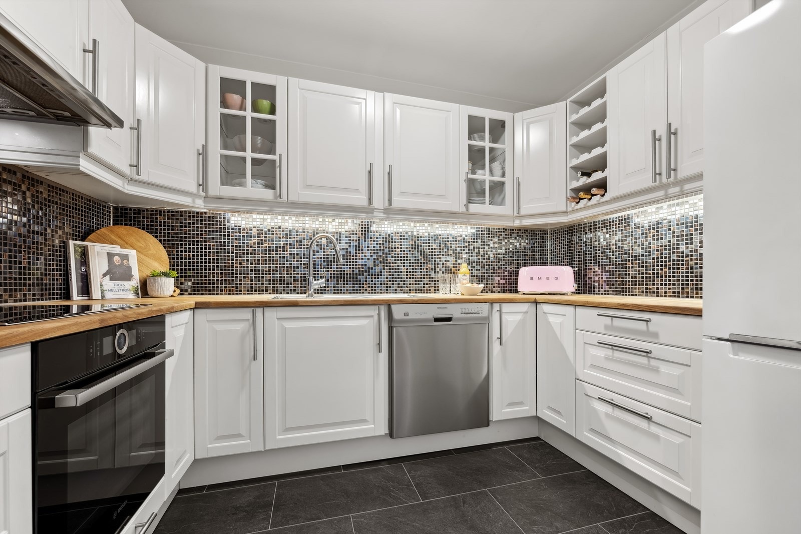 Frontene er lyse og profilerte, med innslag av glass i overskap. Flislagt vegg mellom kjøkkenbenk og overskap er både dekorativt og praktisk for daglig renhold. Galleribilde