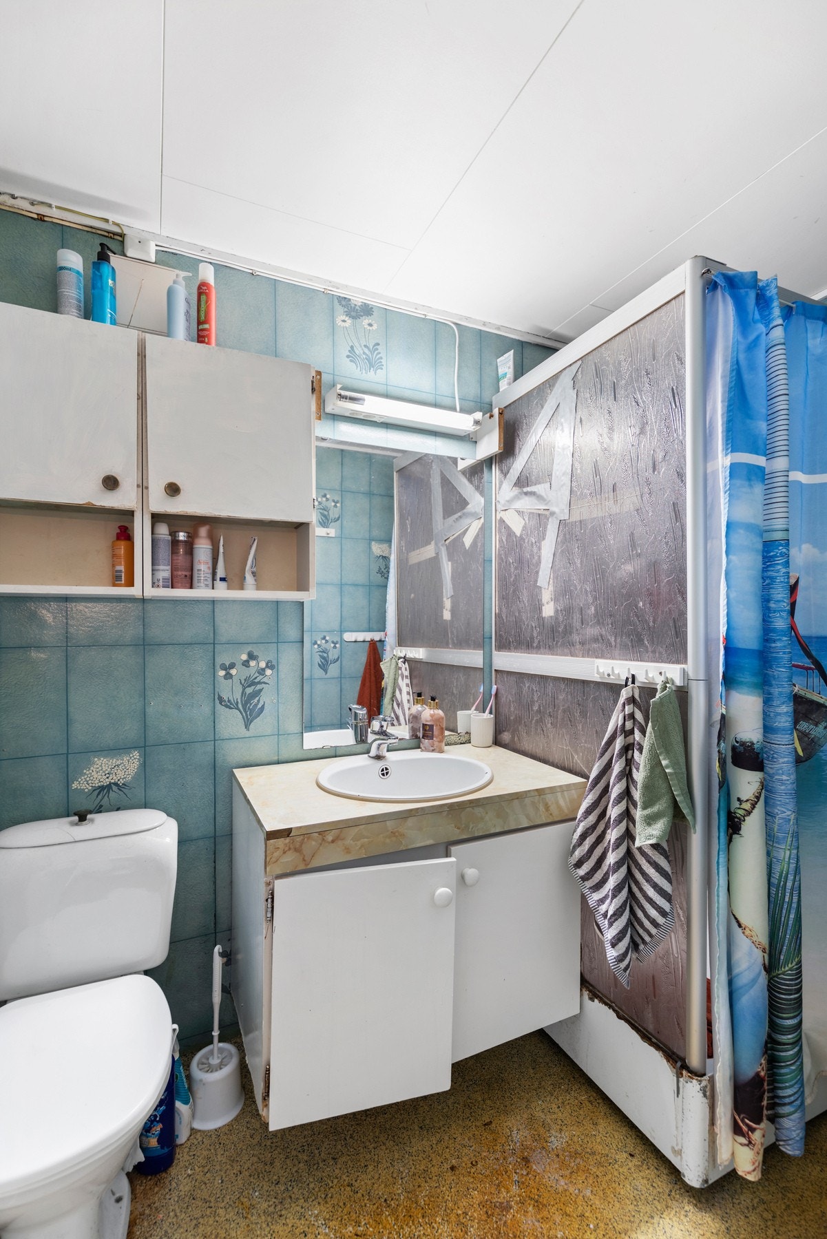 Badet har perginolgulv og tapet på vegger. Rommet er utstyrt med dusjkabinett, toalett og baderomsinnredning med servant og speil. Galleribilde