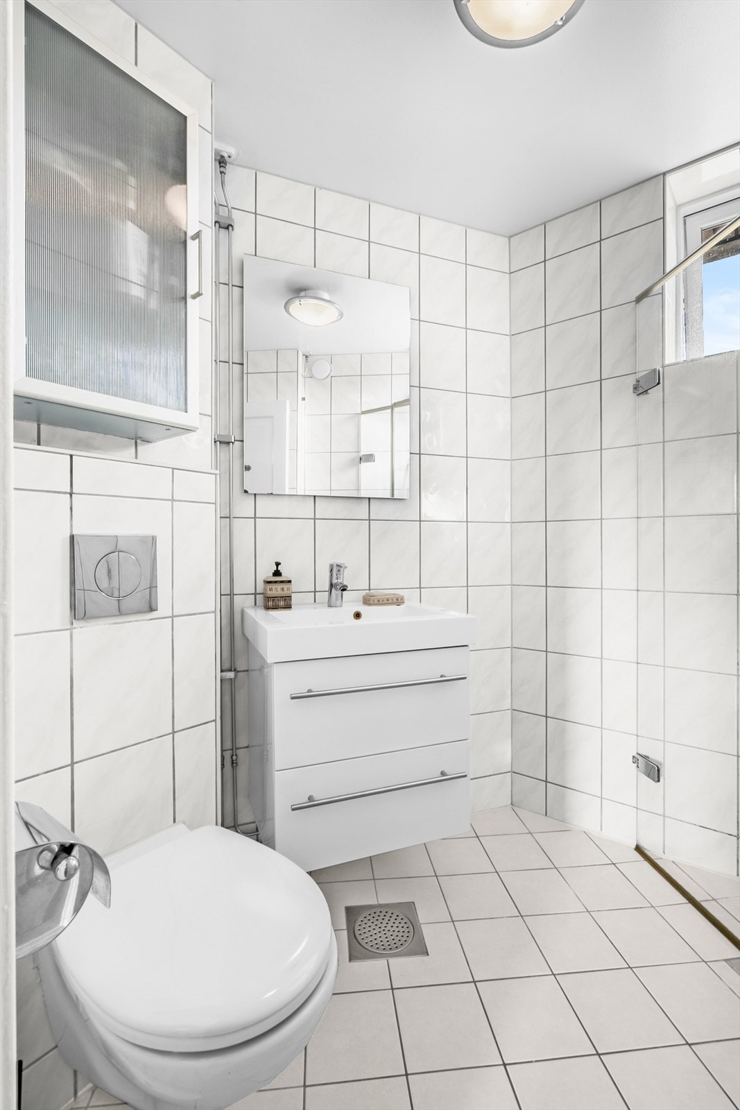 Baderomsinnredning bestående av benkeskap med glatte fronter. Dusj med innfellbare dører i herdet glass, servant, veggfestet klosett med innebygget sisterne og opplegg for vaskemaskin. Galleribilde