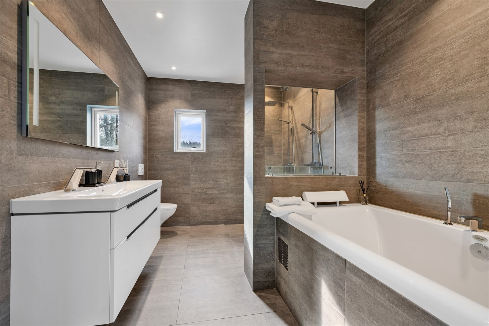 Baderom med dobbel servant, veggmontert toalett, dobbel dusj og innmurt badekar. Herlig biopeis innfelt i vegg og balansert ventilasjon. Galleribilde