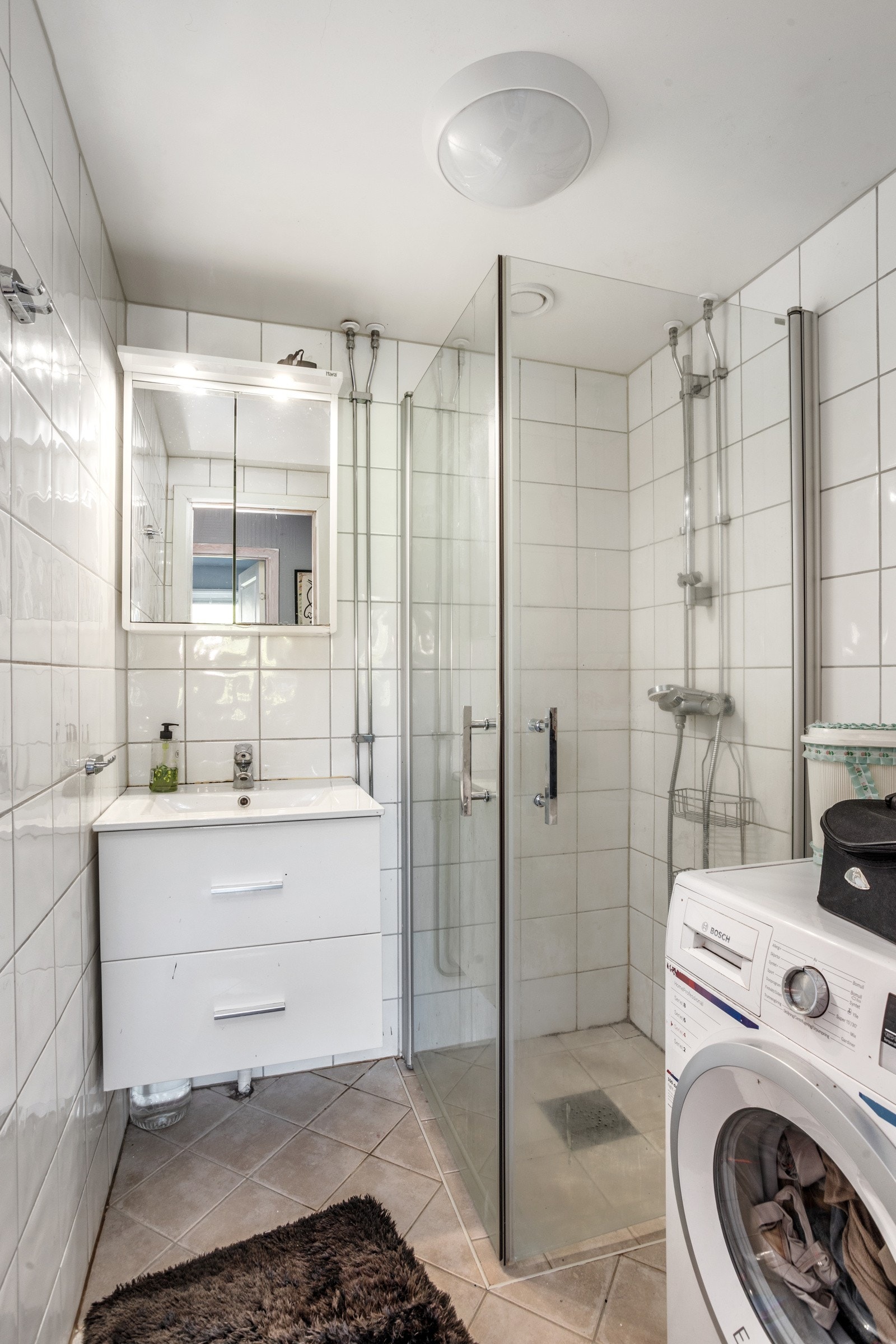 Bad fra 2014, flislagt gulv med gulvvarme og flislagte vegger. Her er det dusjhjørne, opplegg for vaskemaskin, vegghengt servantinnreding med skufffer og speilskap med lys. Galleribilde