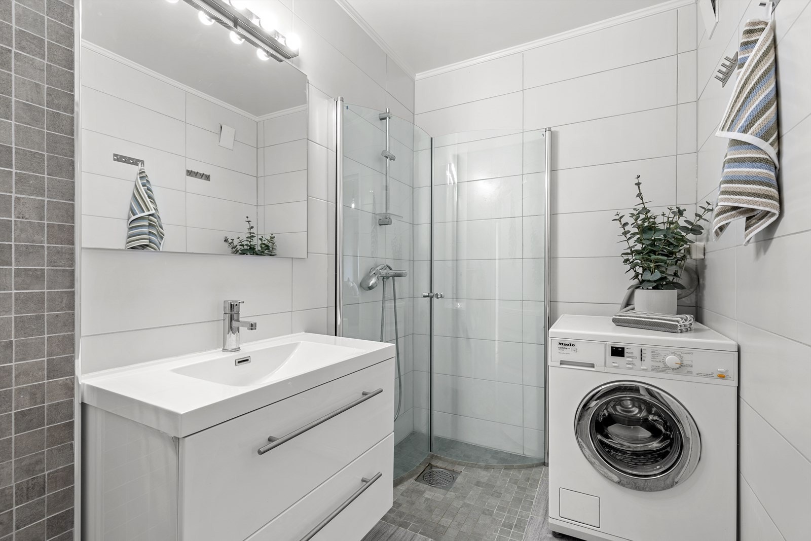 Badet er innredet med nedfelt servant med underskuffer, speil med lys, veggmontert toalett, dusjvegger/hjørne og opplegg for vaskemaskin. Galleribilde