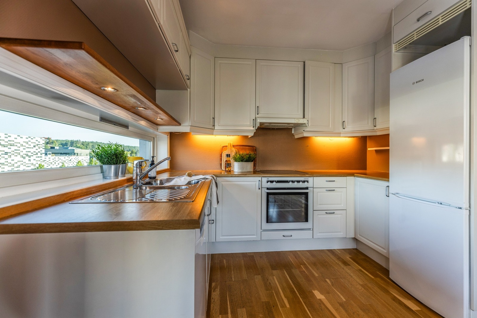 Kjøkkeninnredning med hvite profilerte fronter og benkeplate av laminat. Innredningen har integrert stekeovn, platetopp, og frittstående oppvaskmaskin og kjøleskap. Galleribilde