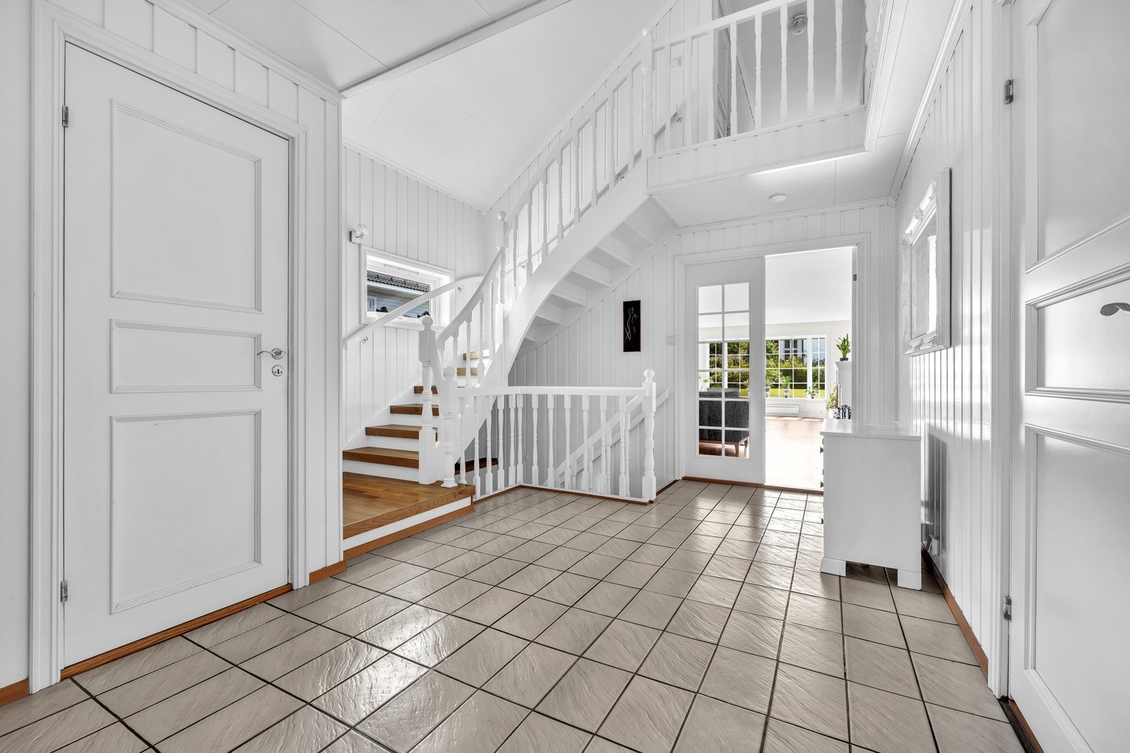 Rommet har fliser på gulvet, hvitmalte flater og trapp opp til andre etasje. Fra hallen er det tofløyet dør med glassfelt videre inn i stuen. Galleribilde
