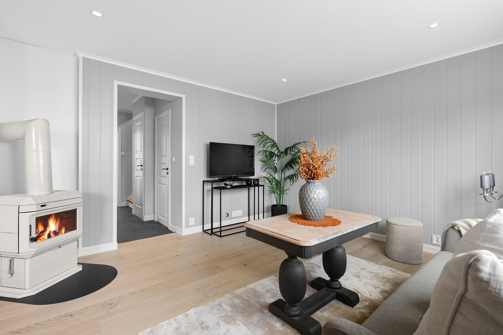 Lys og innbydende stue med trepanel på vegg malt i en fin gråtone, 1-stavs laminat på gulv og utstrakt bruk av downlights i himling. Galleribilde