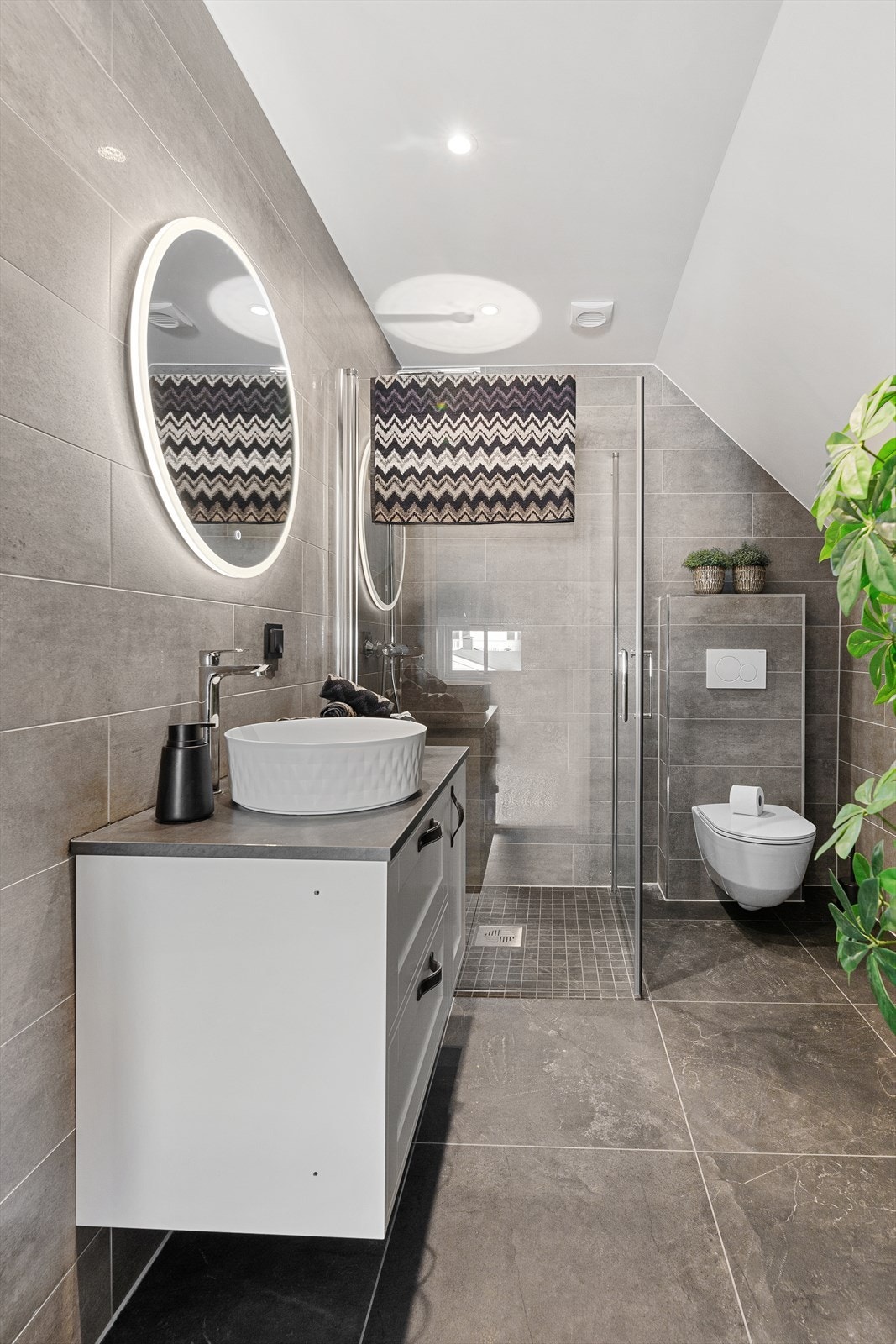 Videre er badet utstyrt med dusjhjørne med mosaikkfliser på gulv, innfellbare dusjvegger og regndusjarmatur i tillegg til vegghengt toalett. Galleribilde