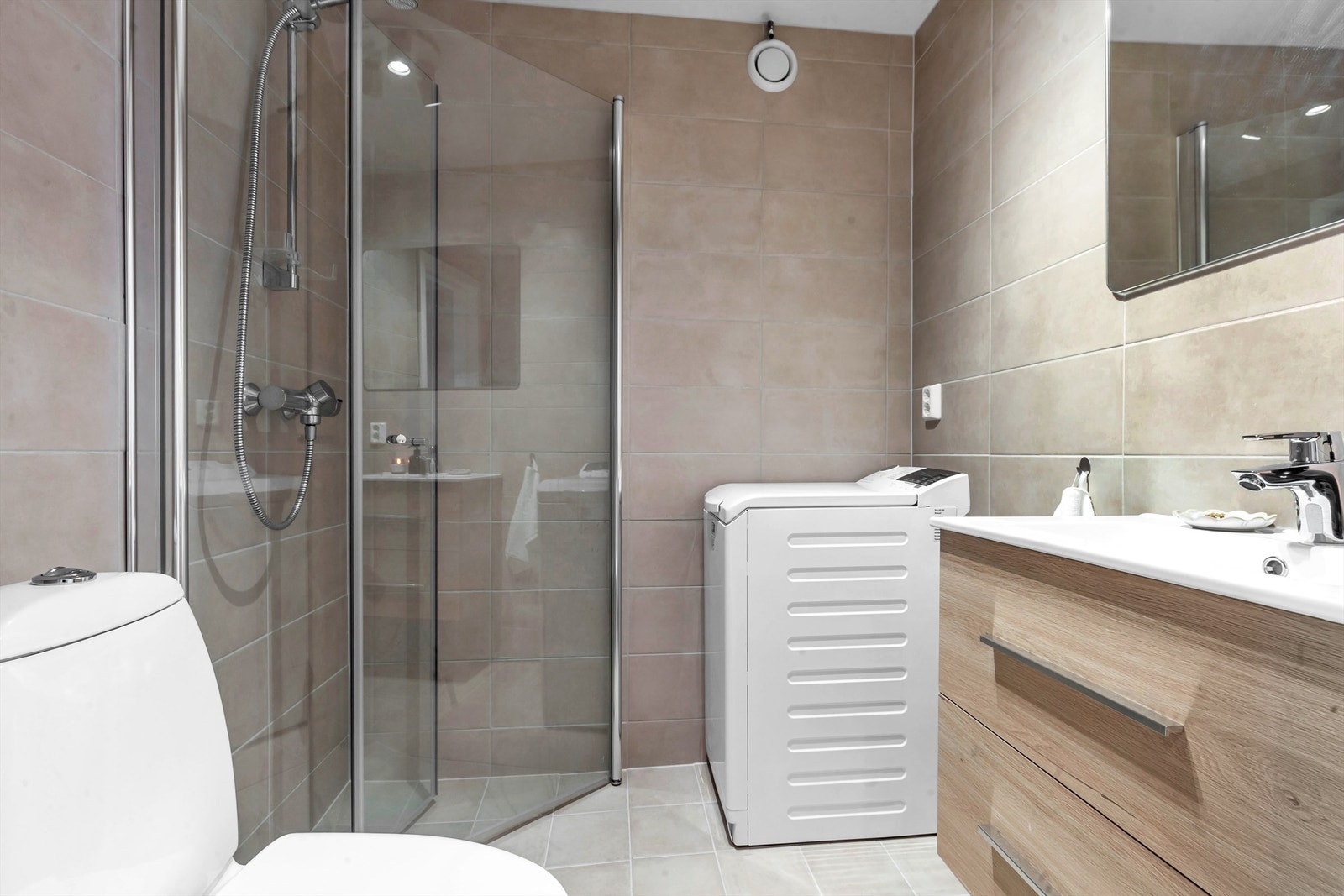 Innbydende flislagt bad med varmekabler i gulv. Dusjhjørne med glassdører, servant med innredning, toalett og opplegg for vaskemaskin. Galleribilde