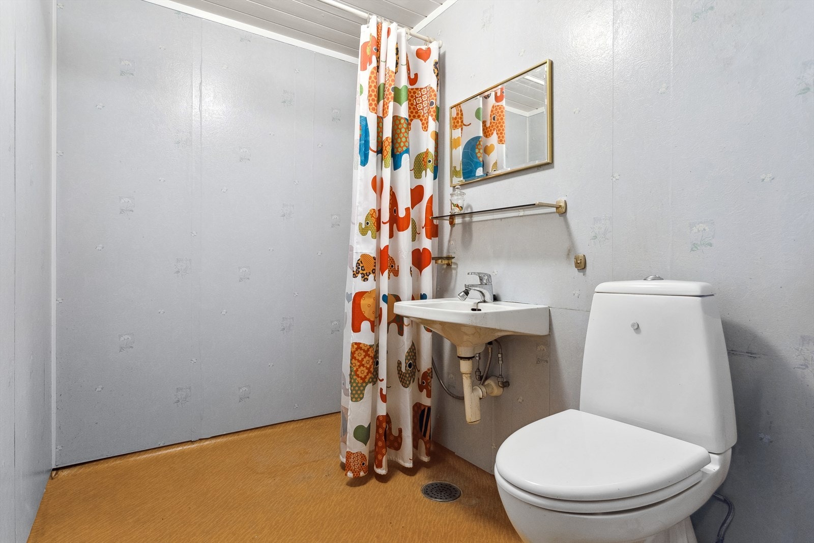 Eldre baderom i underetasjen med belegg på gulv og tapet på vegger. Badet er utstyrt med toalett, servant og dusj med forheng. Galleribilde