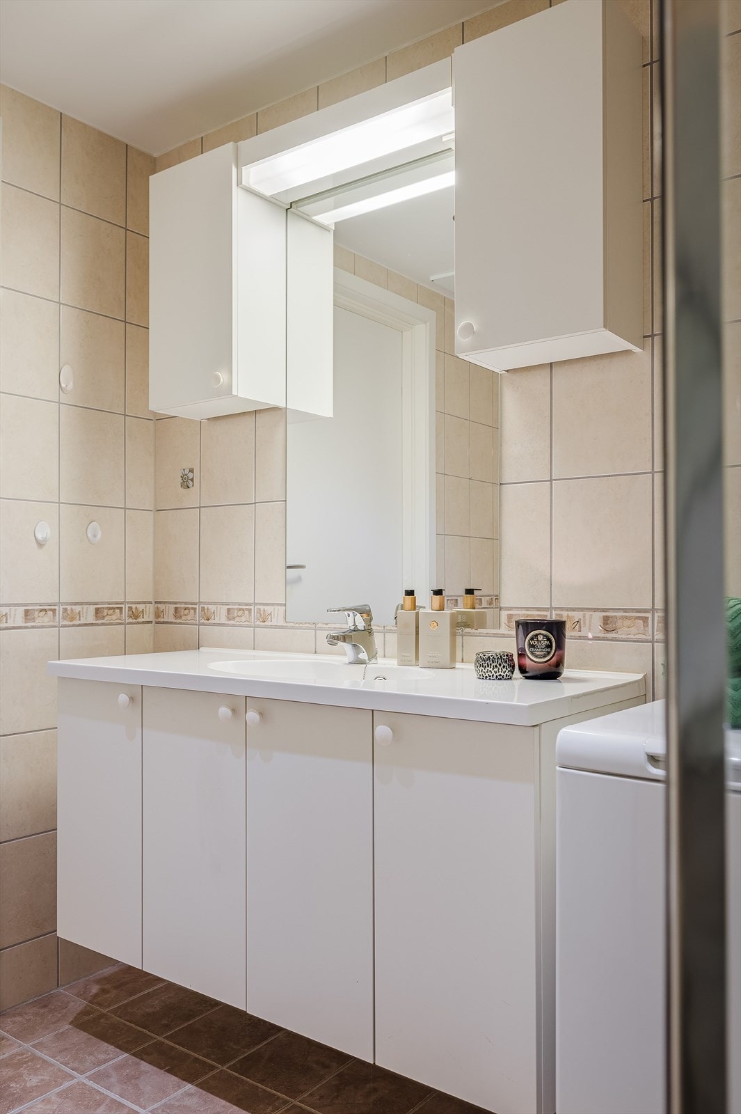 Tidløs baderomsinnredning med lyse fronter og heldekkende servant, speil med belysning og vegghengte skap over. Baderommet er også utstyrt med dusjhjørne med glassdører, toalett og opplegg for vaskemaskin. Galleribilde