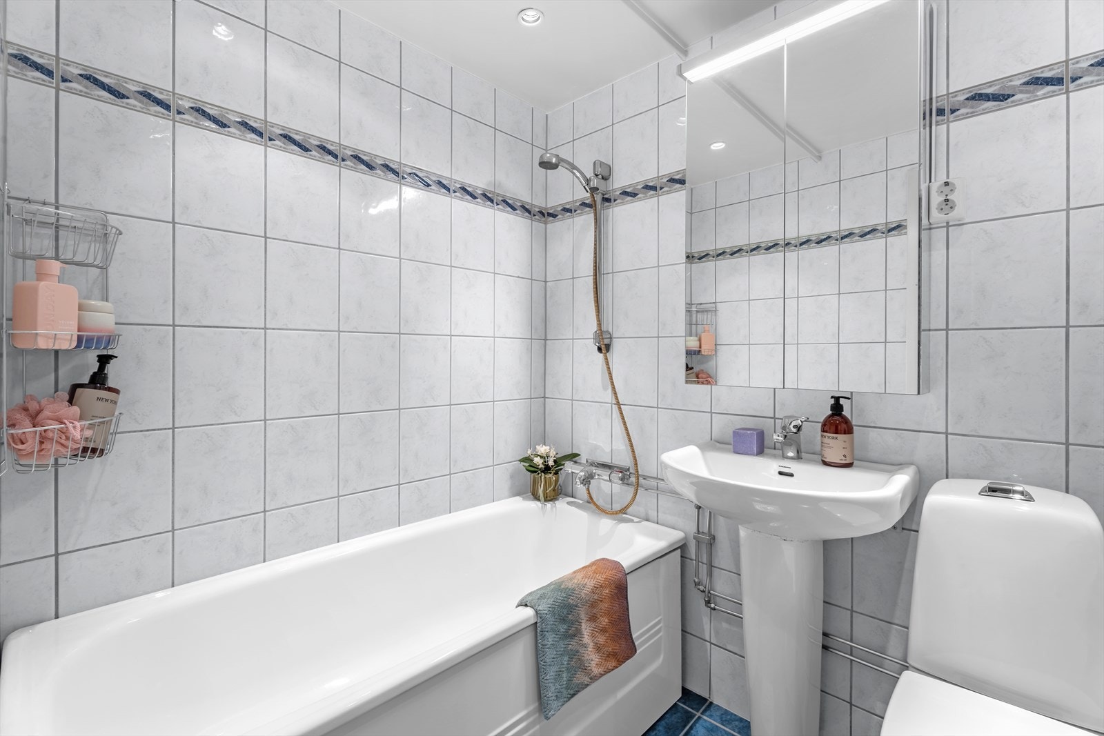 Flislagt bad med behagelig gulvvarme og downlights. Badet inneholder praktisk veggskap med speilfronter og belysning over servant. Dusj i badekar, servant og gulvstående klosett. Galleribilde