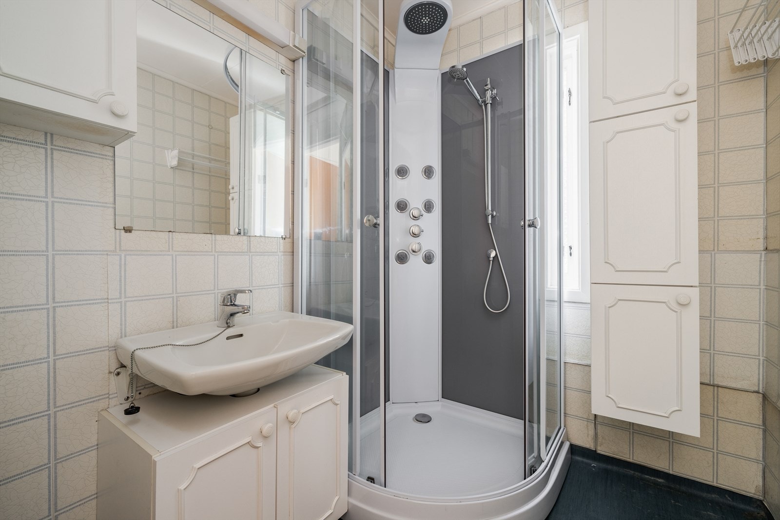 Eldre bad med belegg på gulv, tapet på vegg og takess i tak. Badet inneholder dusjkabinett, servant på vegg med underskap og speil, samt vegghengte skap. Galleribilde