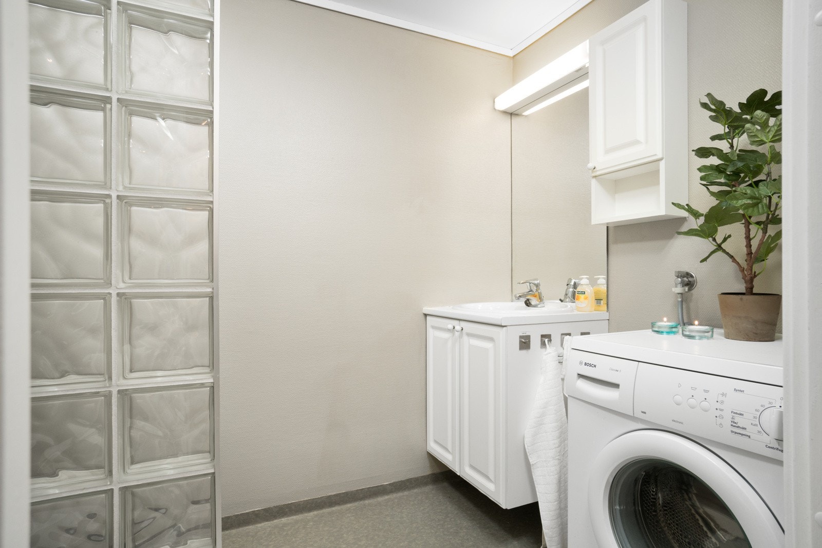 Lyst baderom med praktisk glassbyggervegg. Badet er utstyrt med servant i innredning, toalett og opplegg for vaskemaskin. Galleribilde