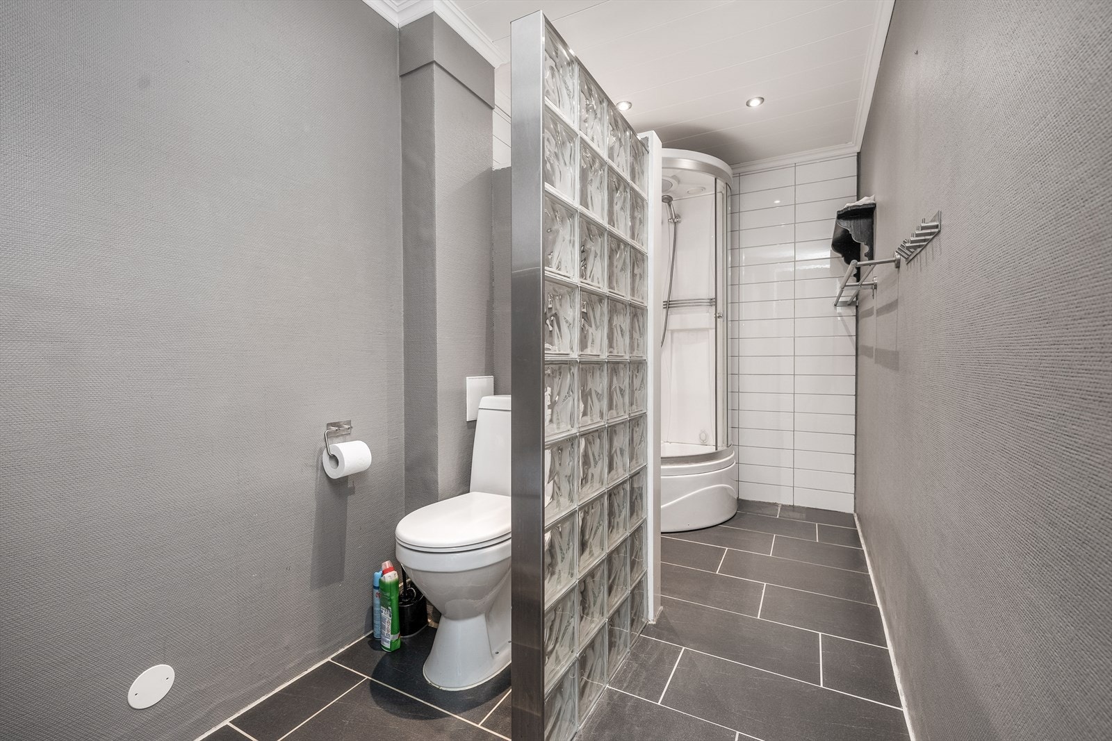 Badet har flis på gulv og på vegg i dusjsone. Toalettet er avskjermet med en vegg av glassbyggersten. Det er satt inn nytt, gulvmontert toalett i 2021. Ellers har rommet en eldre baderomsinnredning i sort fargetone med speil og belysning over vask. Galleribilde