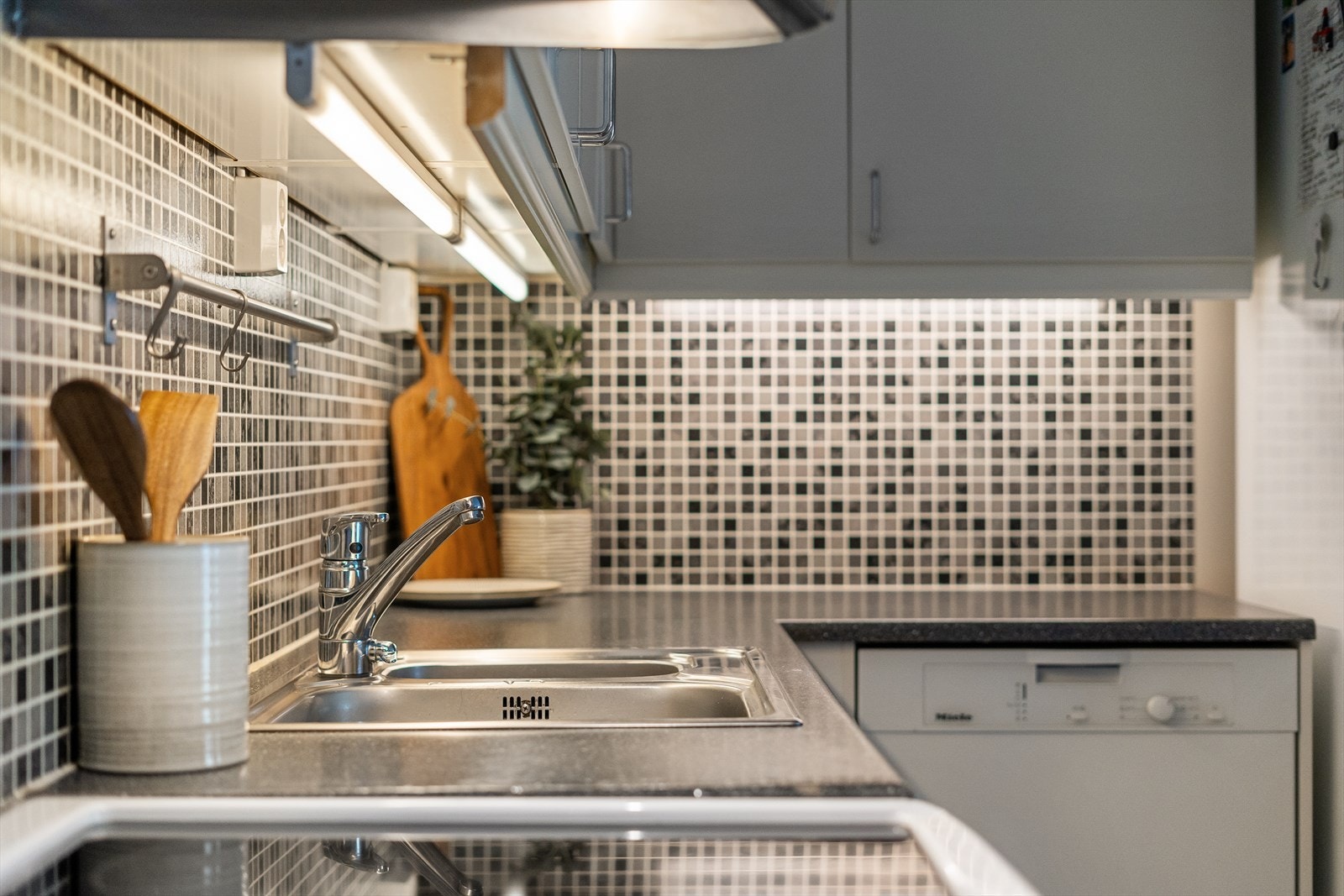 Det er lagt mosaikkfliser på vegg over benkeskap og lyslister og belysning under overskap Galleribilde