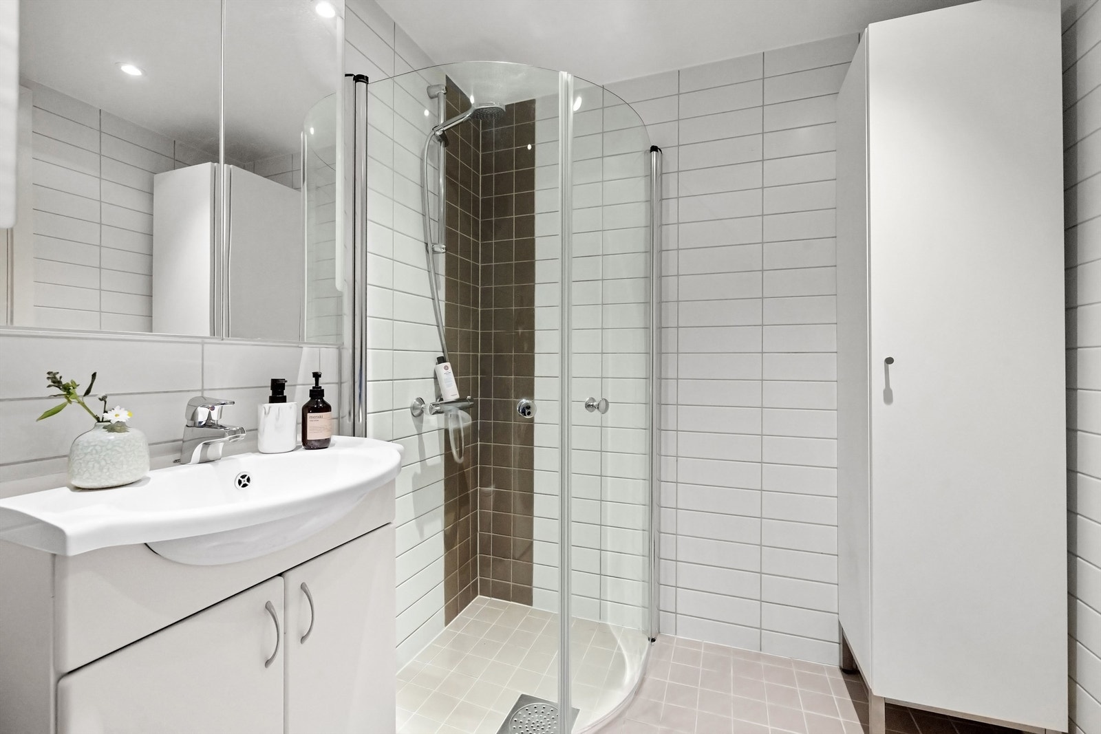 Badet inneholder vegghengt servantinnredning med ett-greps blandebatteri, speilskap på vegg, dusjhjørne med glassdører og vegghengt toalett. Galleribilde