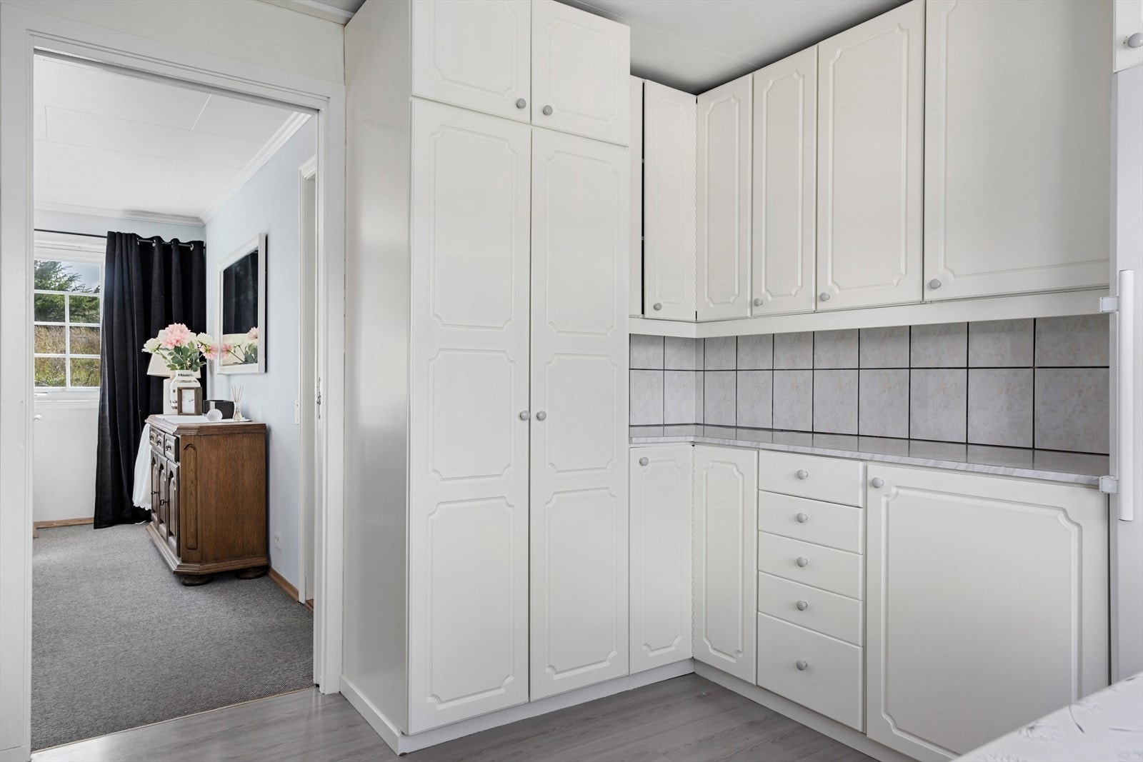 Kjøkken med profilerte fronter og benkeplate av laminat med nedfelt vask og oppvaskbrett i rustfritt stål. Det er fliser mellom benkeplate og veggskap. Hvitevarer medfølger handelen om ønskelig. Galleribilde