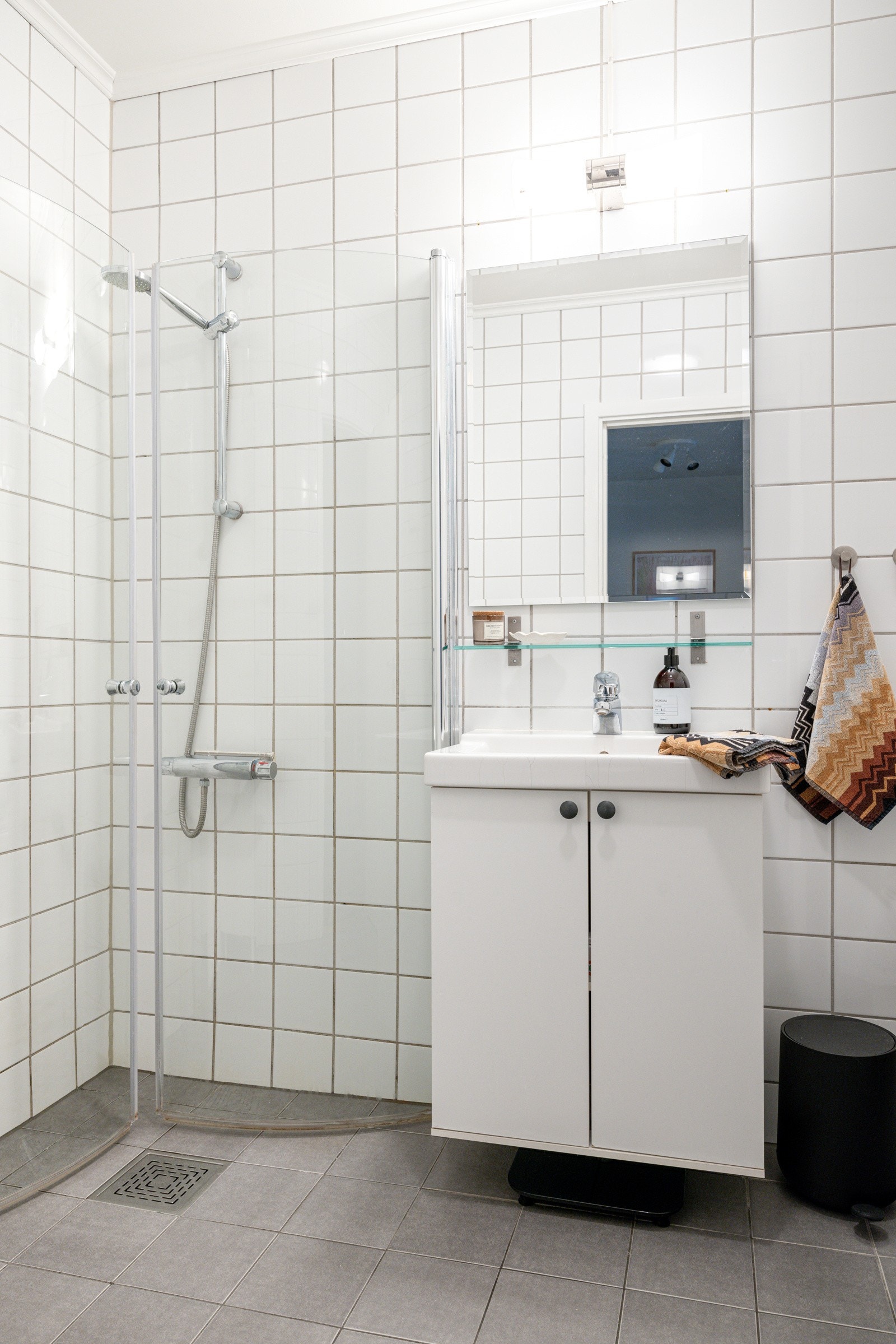 Tidløs baderomsinnredning med ovenpåliggende servant, vegghengt glasshylle og speil med belysning over. Baderommet er også utstyrt med dusjhjørne med innfellbare glassdører, ekstra høyskap for oppbevaring og gulvstående toalett. Galleribilde