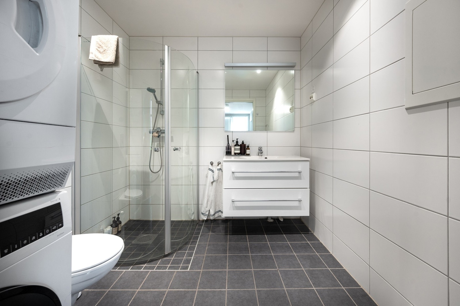 Baderomsinnredning med servantskap i hvit utførelse, dusjvegg og vegghengt wc. Vegger og gulv er belagt med keramiske fliser. Gulvvarme. Galleribilde