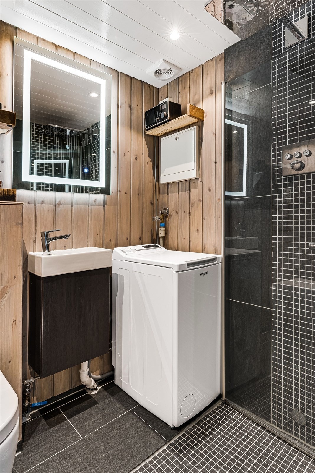 Badet er innredet med servant som er nedfelt i innredning, et flott og belyst speil, god hylleplass, dusjhjørne mned innfellbare dører i glass og vegghengt toalett. Vaskemaskin som medfølger, og ventilasjon via elektrisk vifte i himling. Galleribilde