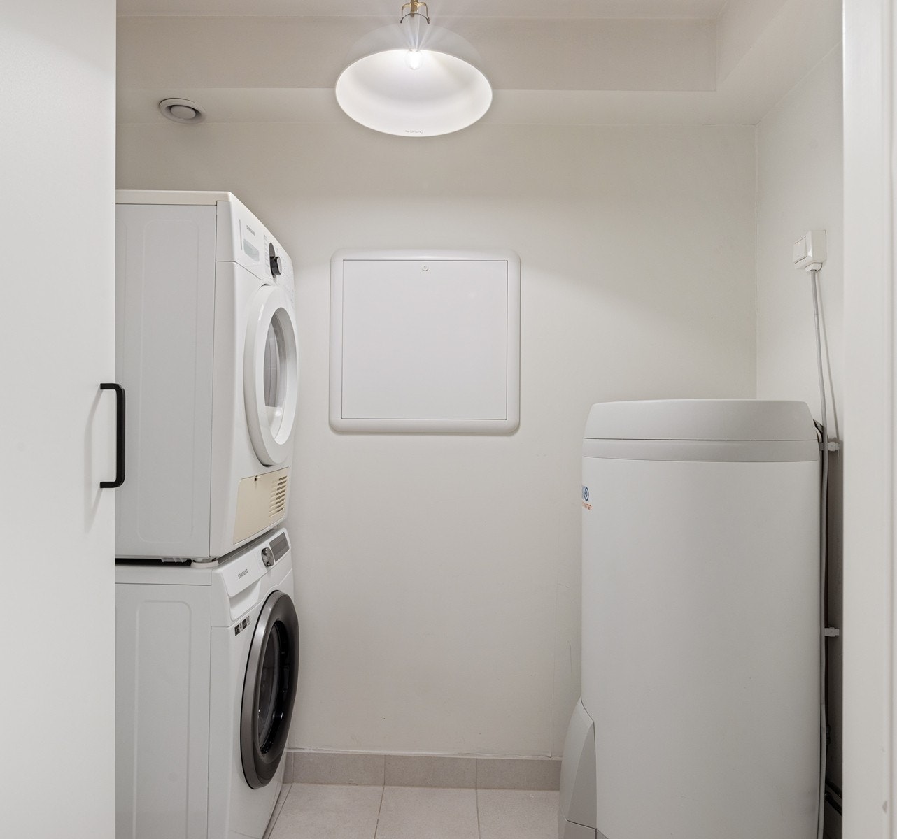 Praktisk vaskerom med flislagt gulv med gulvvarme og malte glatte flater på vegger. Innredningen består av et sideskap og det er opplegg for vaskemaskin og tørketrommel. Galleribilde