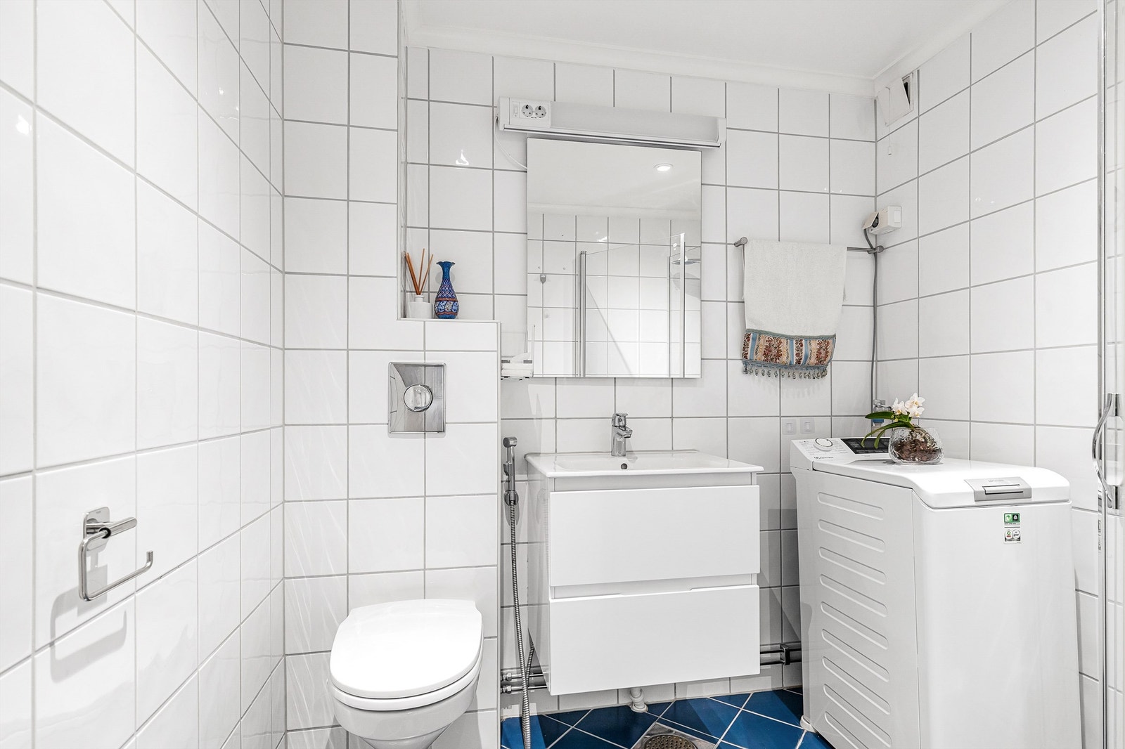 Badet er pent flislagt på gulv og vegger, innredet med vegghengt toalett, vegghengt servantinnredning med heldekkende servant og speil på vegg med overlys. Galleribilde