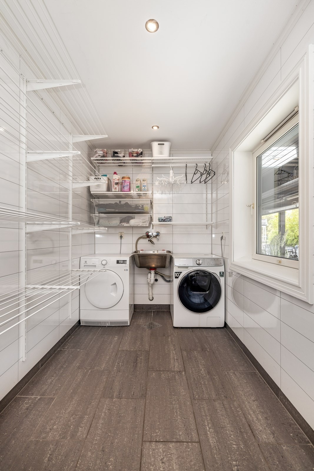 Vaskerommet er praktisk innredet med hyller, opplegg for og plass til vaskemaskin og tørketrommel, samt utslagsvask. Overflater er fliser på gulv med varmekabler og baderomspanel på vegger. Downlight belysning i tak. Galleribilde