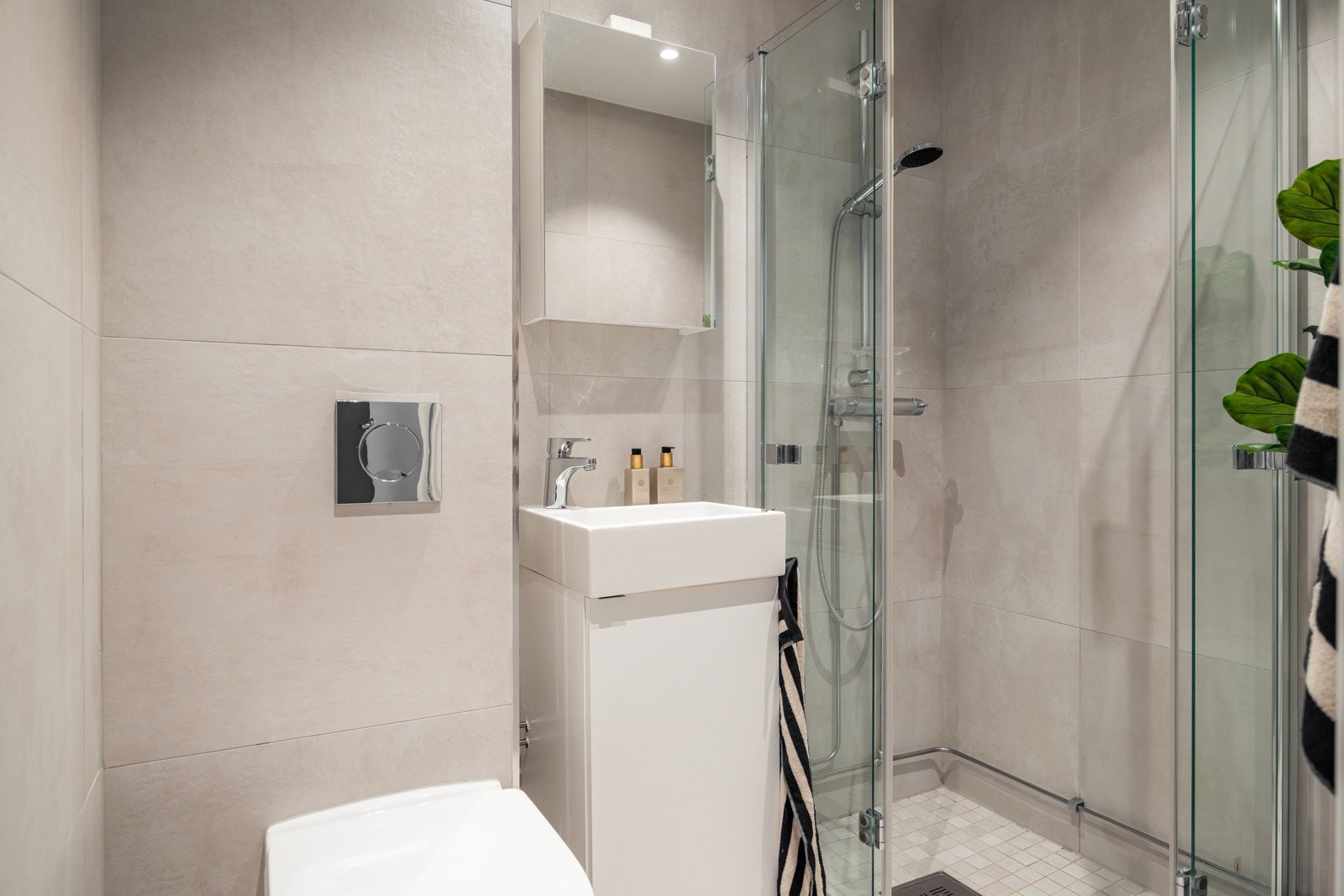 Moderne baderomsinnredning med skuffer og heldekkende servant, speil med belysning over. Baderommet er også utstyrt med dusjhjørne med nedsenk, innfellbare dusjvegger, veggmontert toalett og opplegg for vaskemaskin. Galleribilde