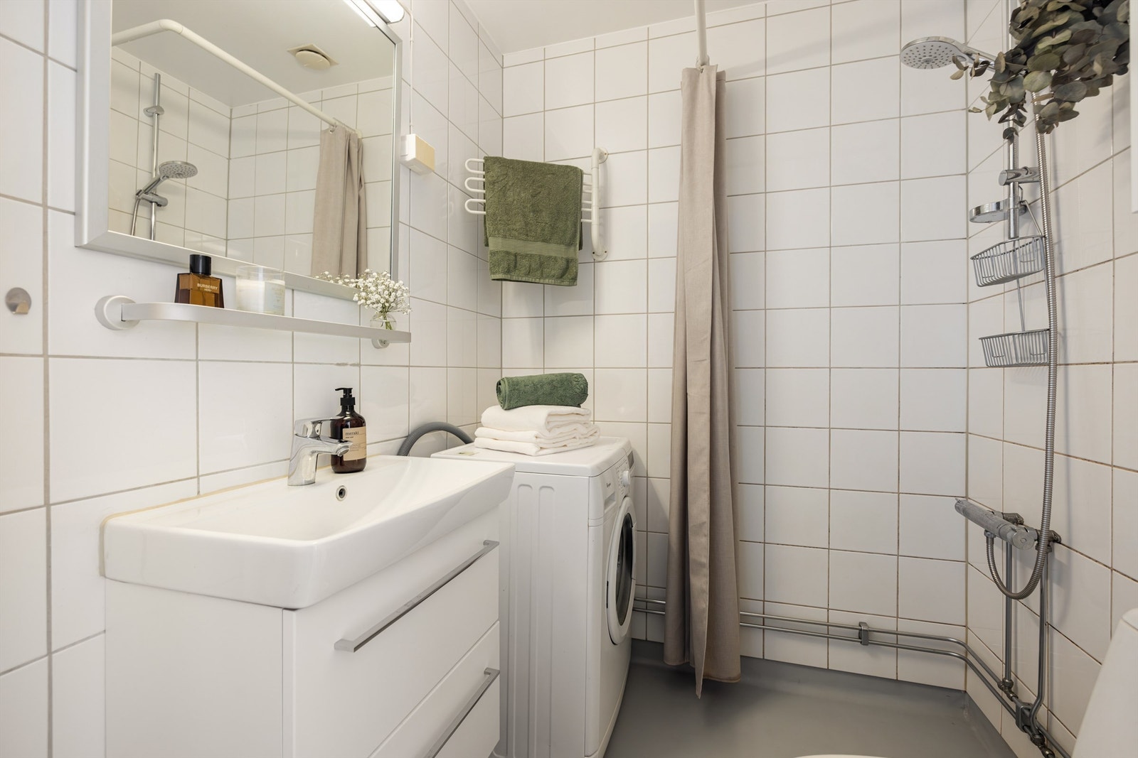 Baderom innredet med vegghengt servantskap m/ovenpåliggende servant, speil m/overlys og stikkontakt på vegg, dusjhjørne, gulvstående toalett og opplegg for vaskemaskin. Overflater; gulvbelegg, flislagte/malte veggflater, himling av malt mur. Galleribilde
